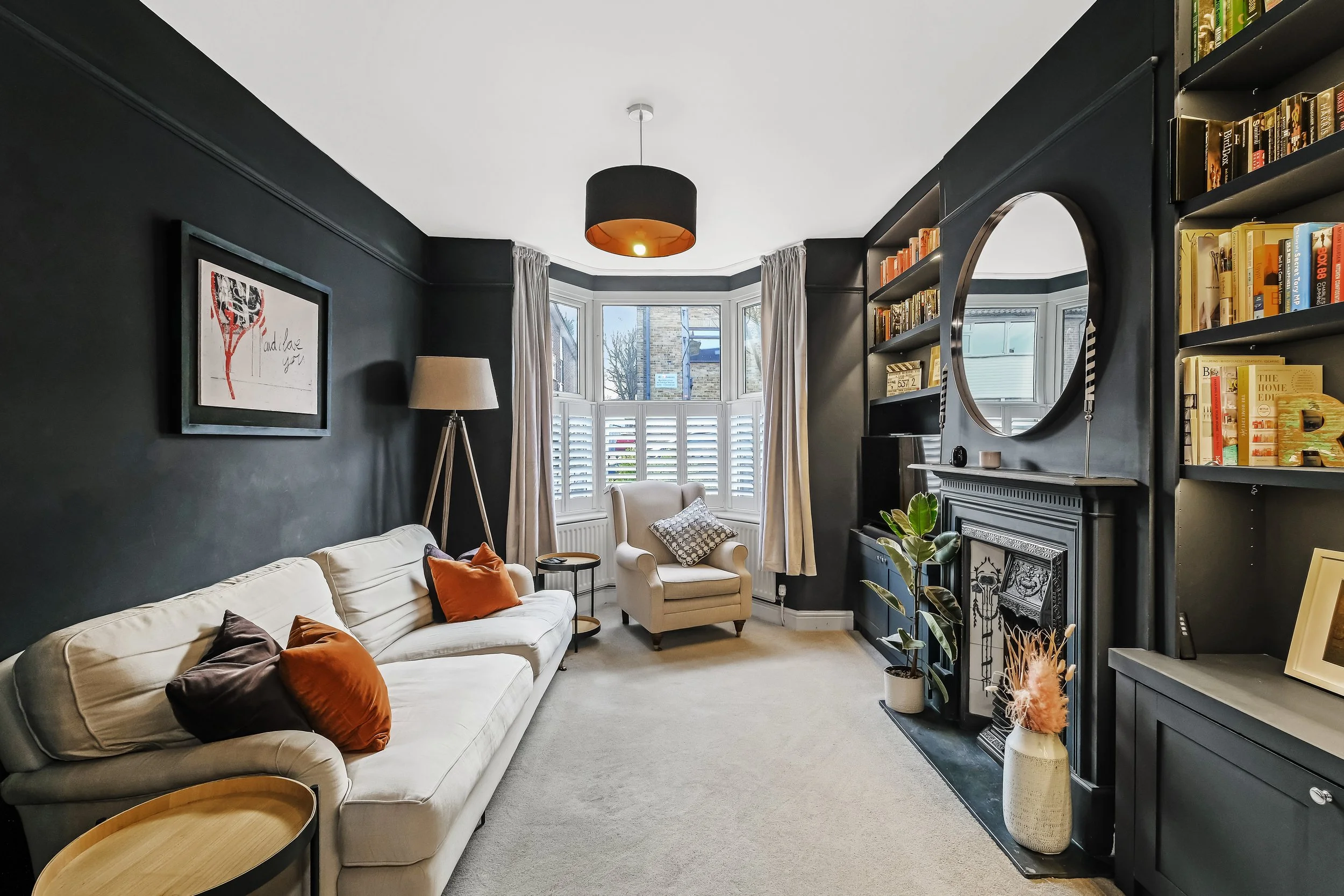 Living room with dark walls, beige sofa with colorful pillows, armchair near bay window, black fireplace with mirror, bookshelves, potted plant, and artwork on wall.