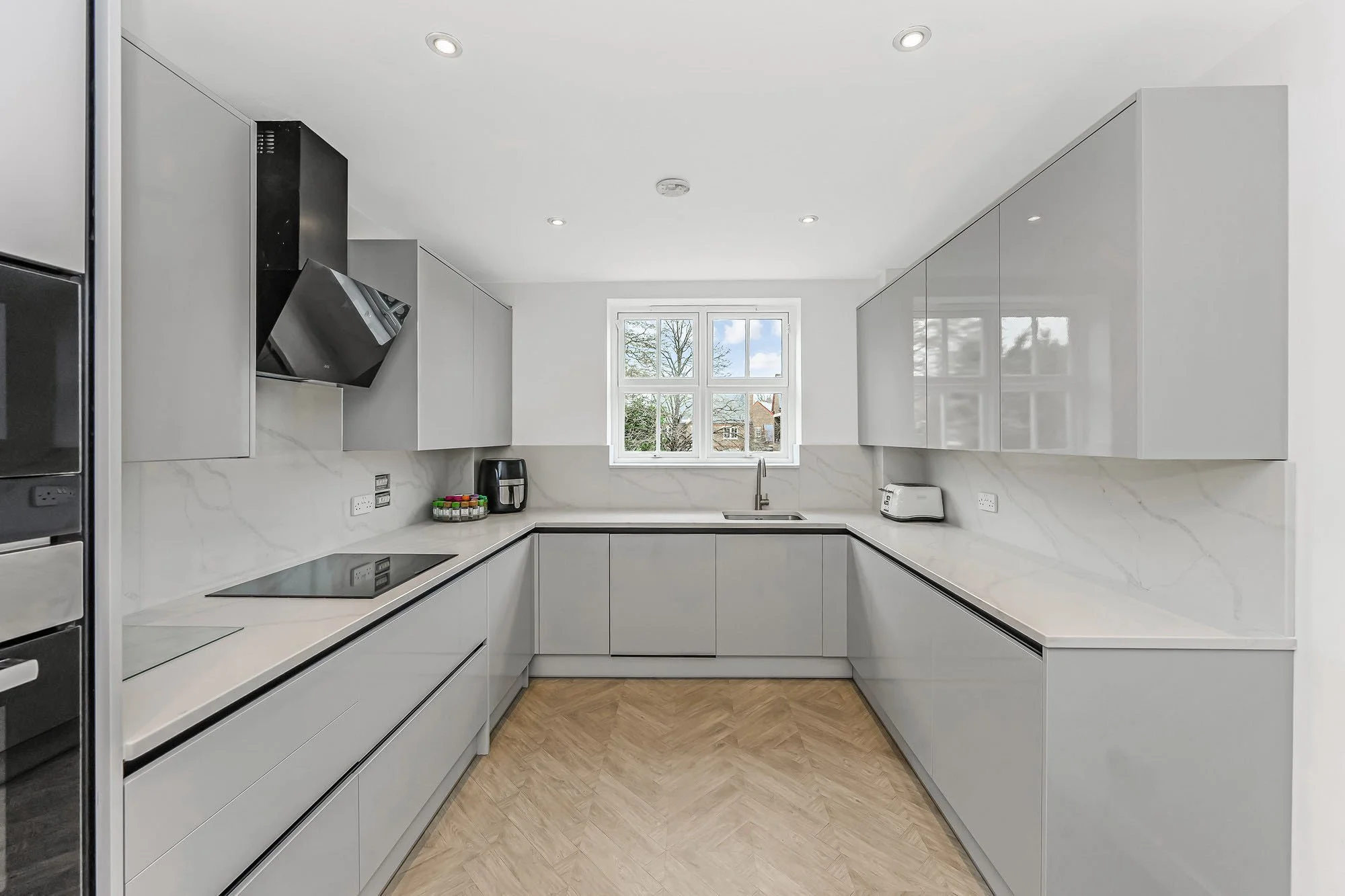 Modern U-shaped kitchen with white cabinetry, marble countertops, a window overlooking trees, and various small appliances on the counter.