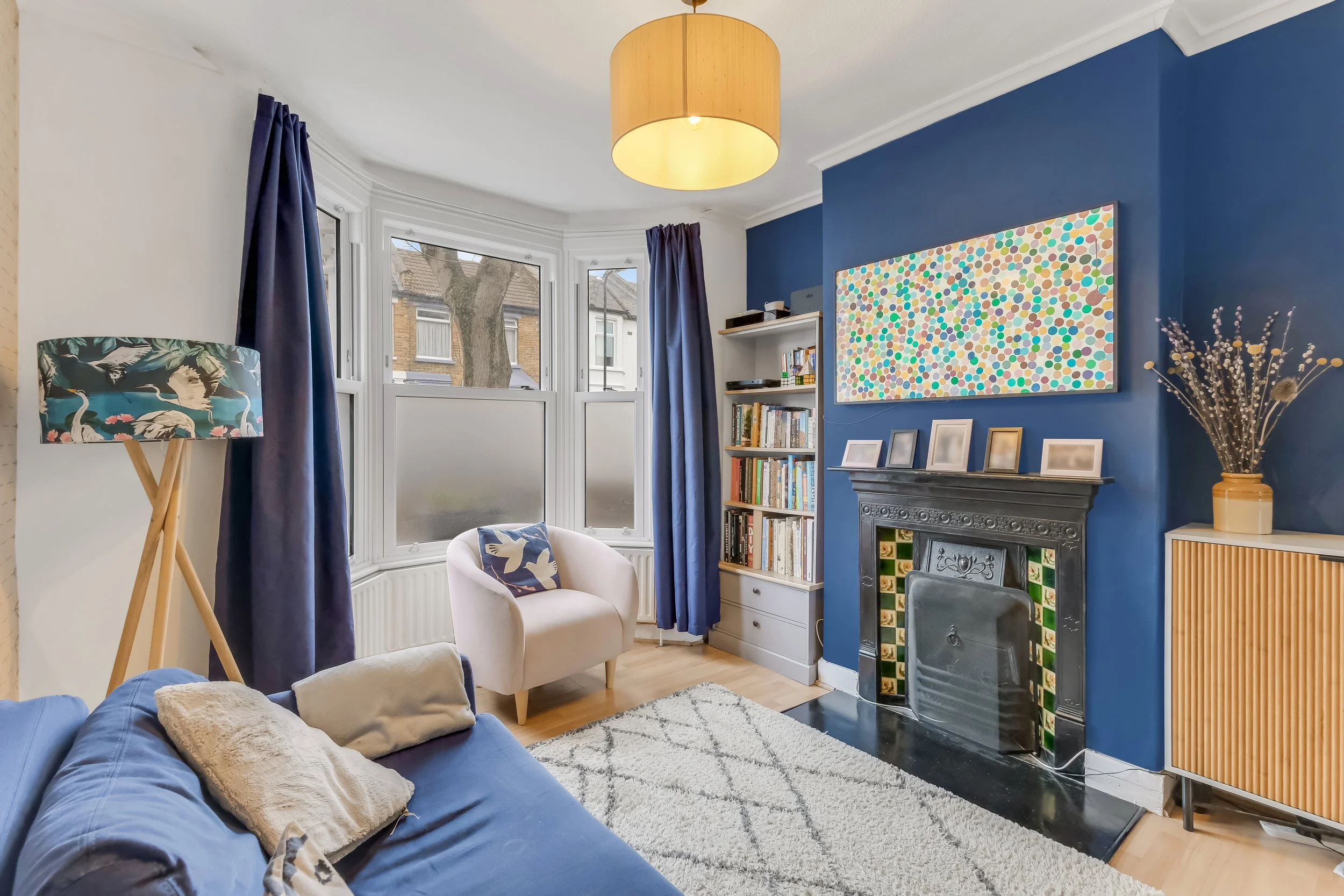 A living room with a blue accent wall, fireplace with decorative tiles, white bookshelf filled with books, a beige armchair with a patterned pillow, a blue sofa with cushions, a white rug, a floor lamp with a patterned shade, large bay windows with w