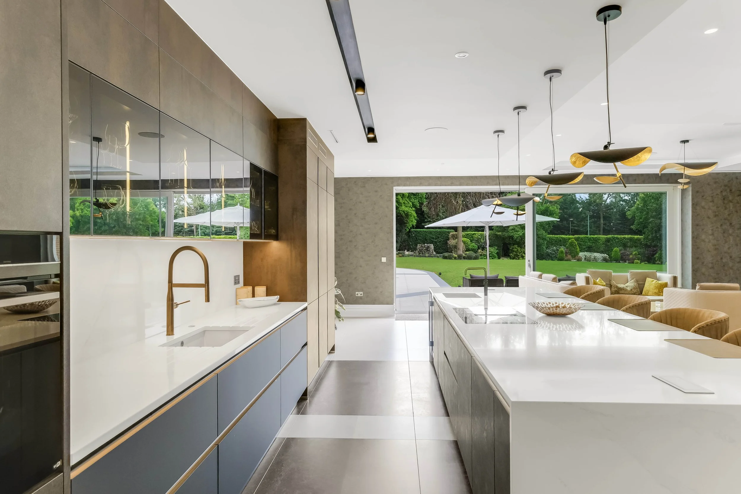 Modern kitchen with blue lower cabinets, white countertops, a brass faucet, and a view of a lush green backyard with outdoor furniture and umbrellas.