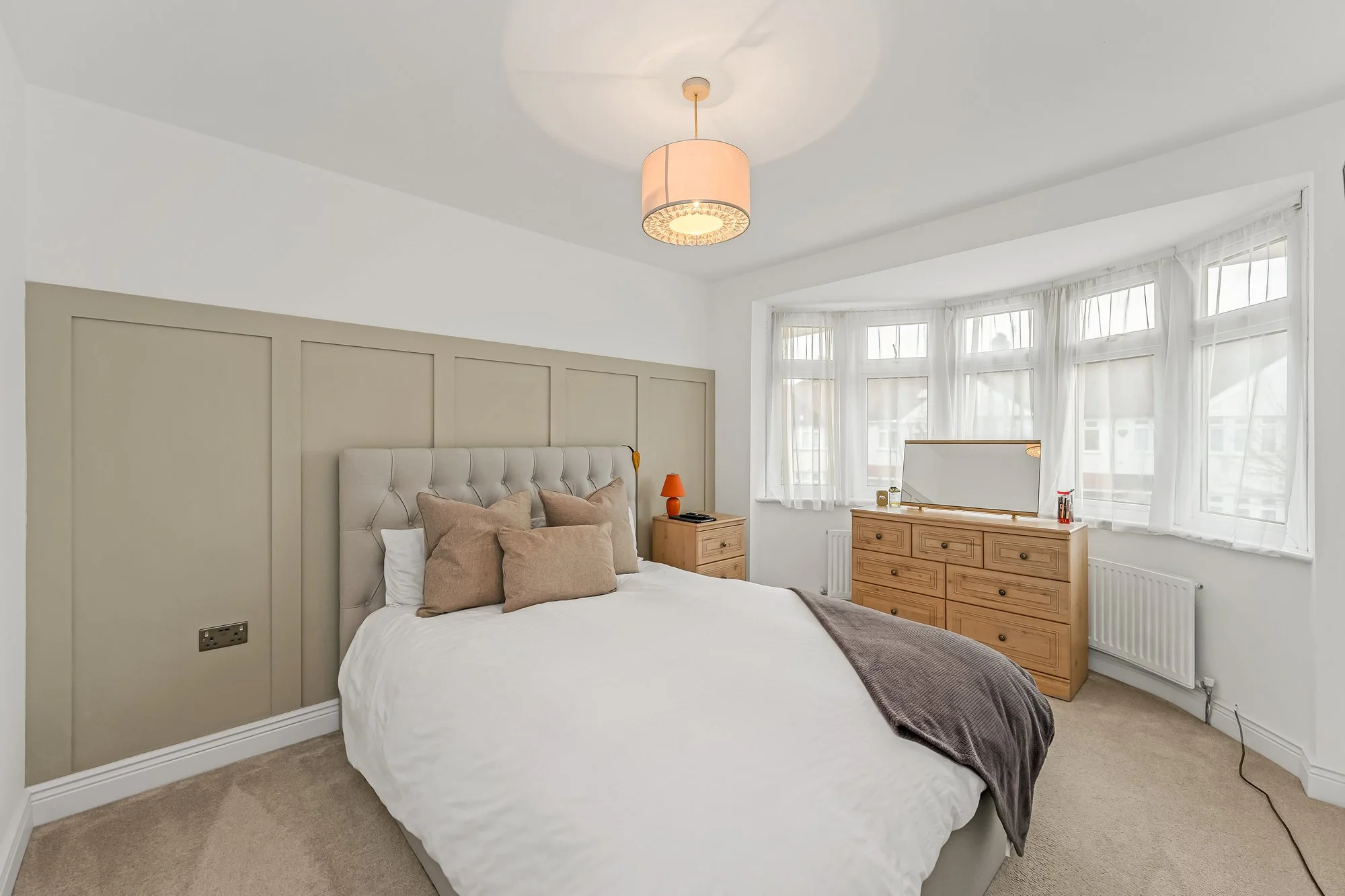 A bedroom with a large bed with a beige headboard, brown and beige pillows, and a brown throw blanket. There is a wooden dresser with a mirror on top, and a small orange lamp on a nightstand. The room has white walls, a beige carpet, and large window