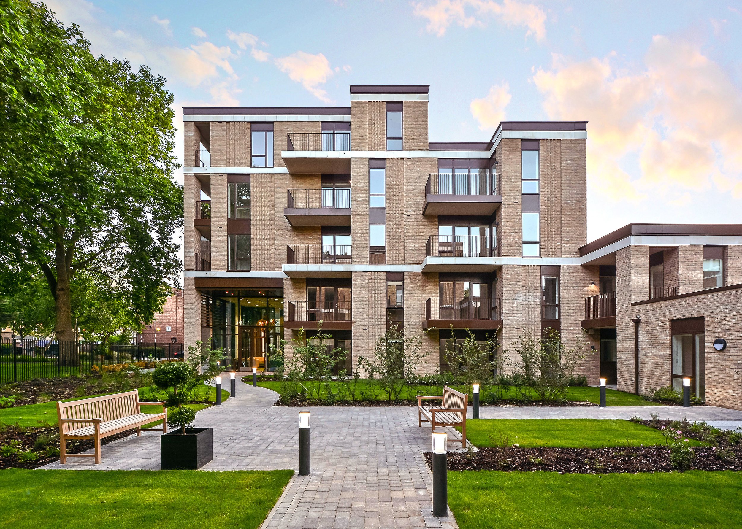 Modern multi-story apartment building with balconies and large windows, surrounded by a landscaped courtyard featuring benches, small trees, and pathway lighting, under a partly cloudy sky.