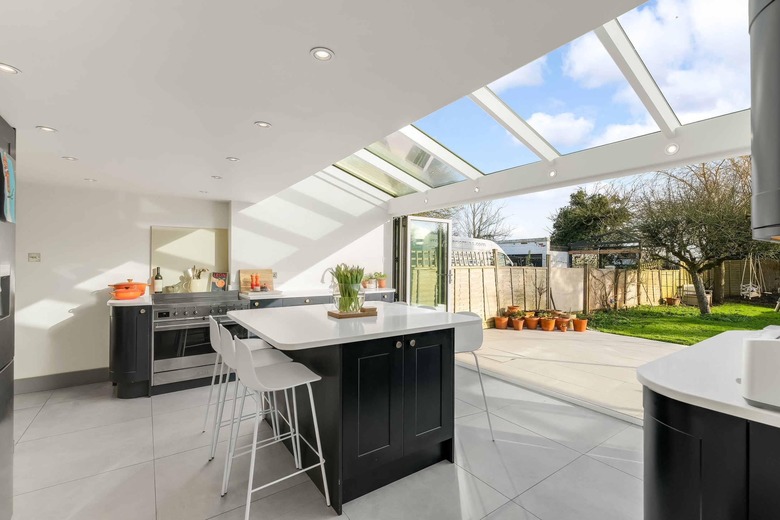 Modern kitchen with white walls, black cabinets, and large glass doors leading outside. Skylights in the ceiling provide natural light. Outside are potted plants, a lawn, and trees.
