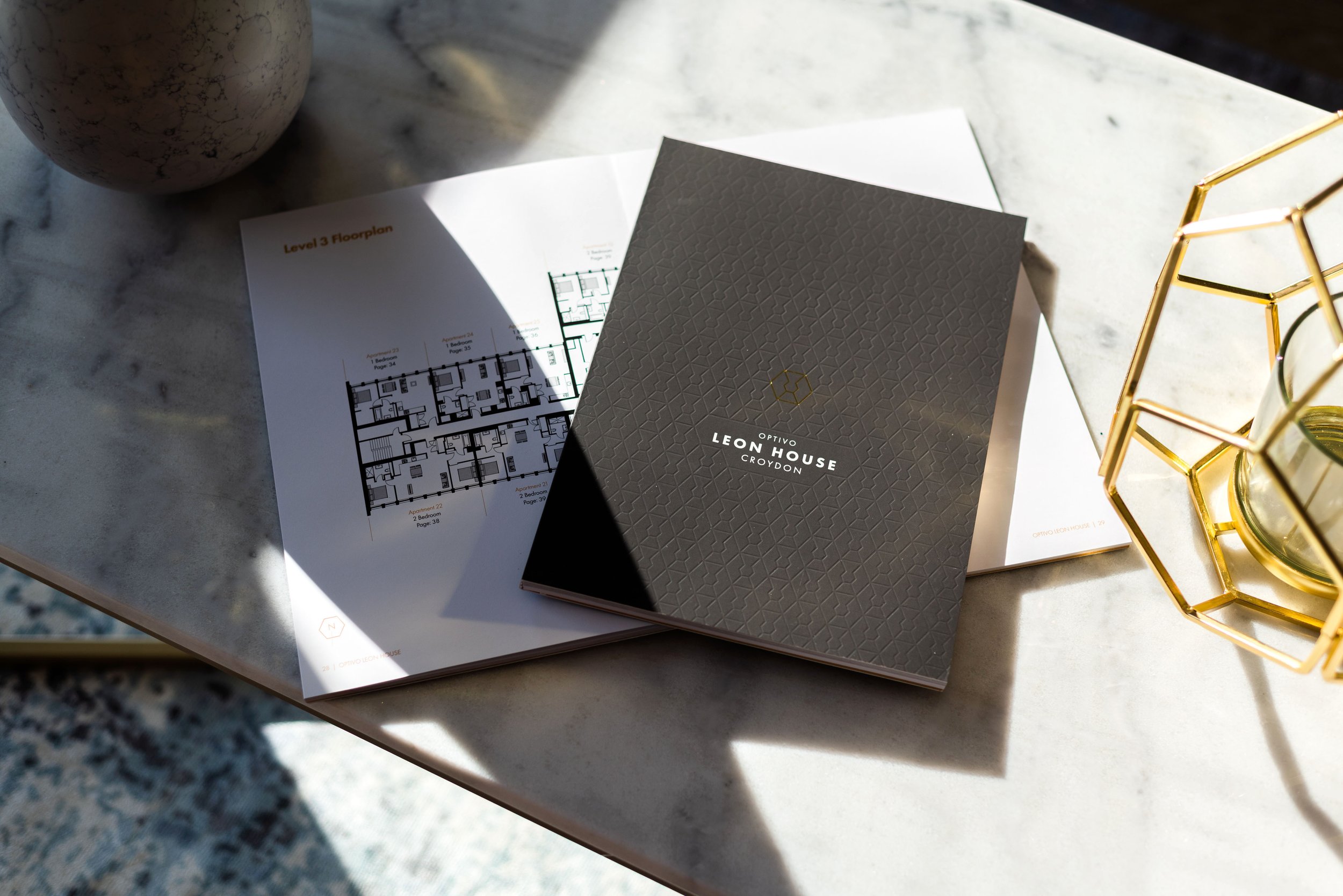 A marble tabletop with a gray booklet titled "Optivo Leon House Croydon," a floor plan sheet, and a gold geometric candle holder with an unlit candle. The scene is lit by natural light, creating shadows.