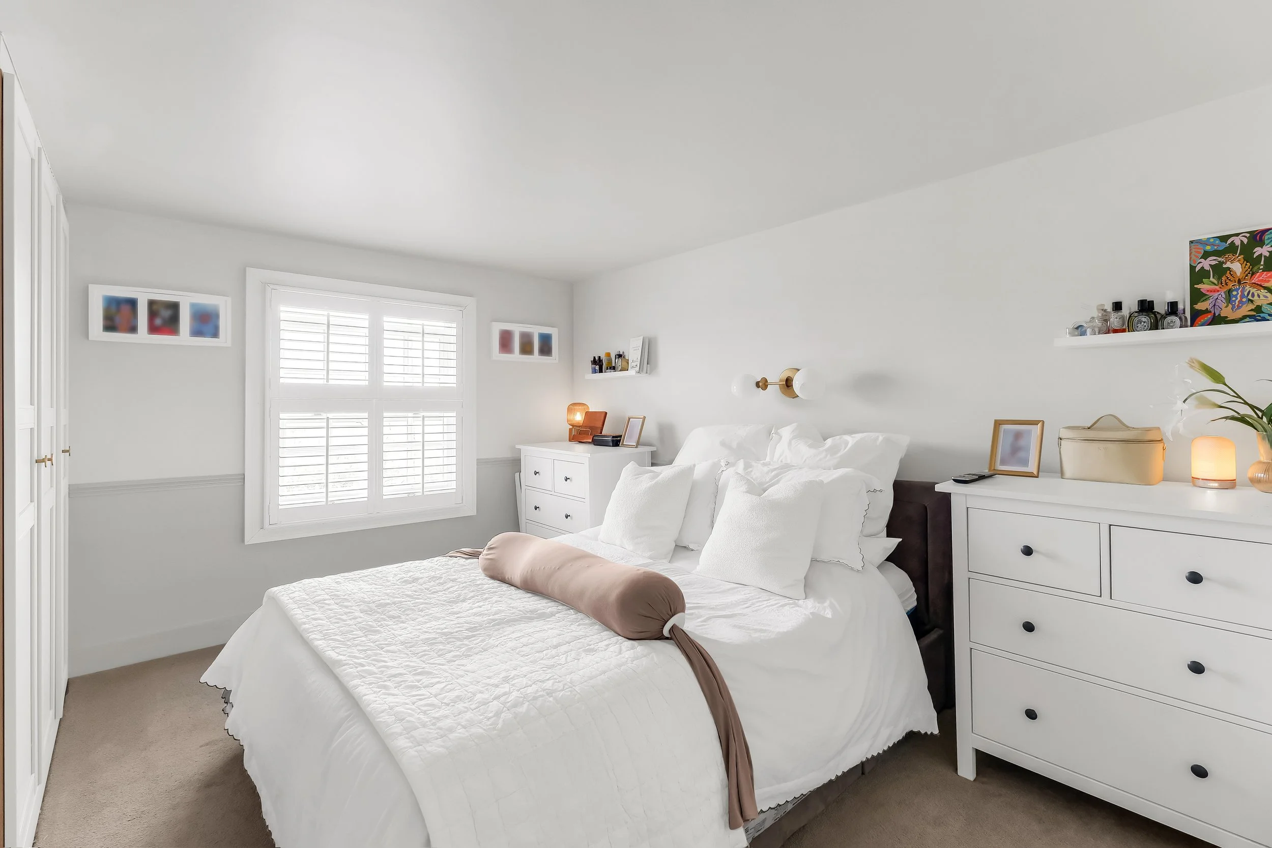 A bright bedroom with white walls, a bed with white linens and a beige bolster pillow, two white dressers, and wall-mounted light fixtures. There are framed artworks on the walls and small decorative items on the dressers and shelves.