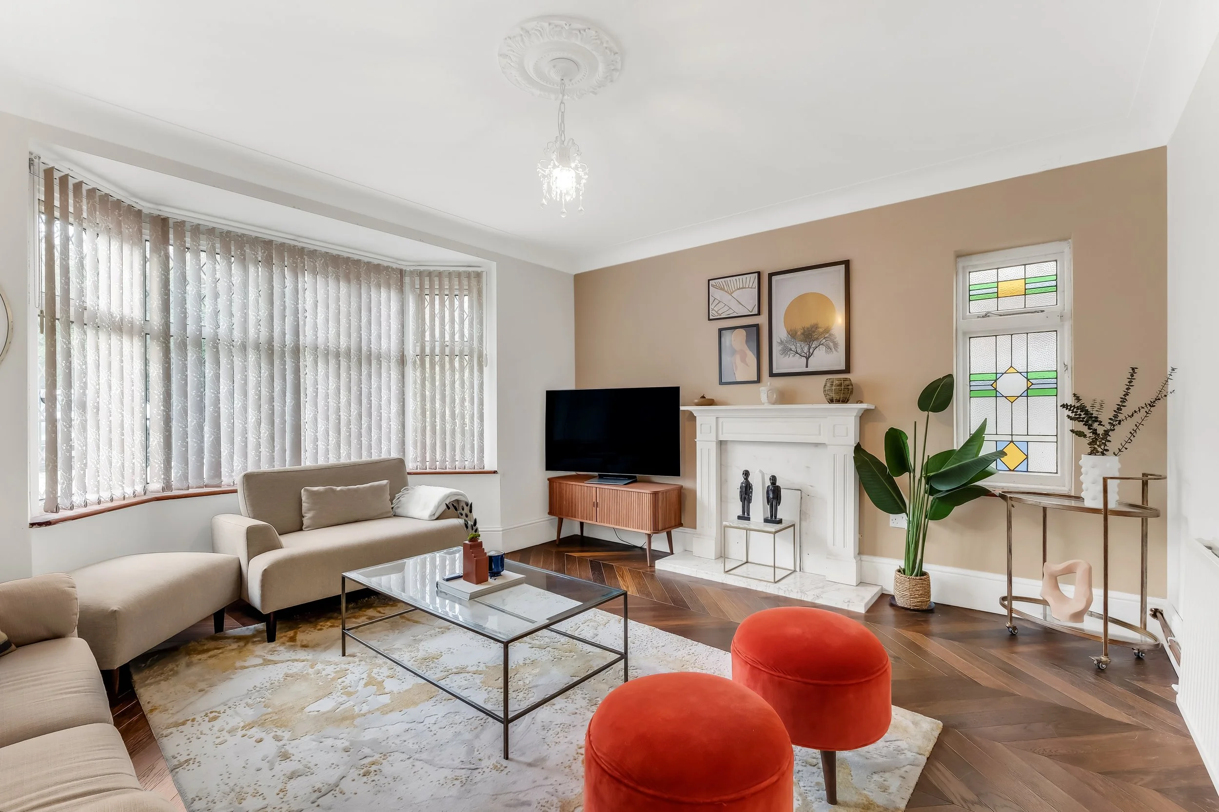 A cozy living room with beige sofas, a glass coffee table, a white fireplace mantel with decorative objects, framed artwork, stained glass windows, a flat-screen TV, lush green houseplants, a gold side table, and a ceiling chandelier, with hardwood f