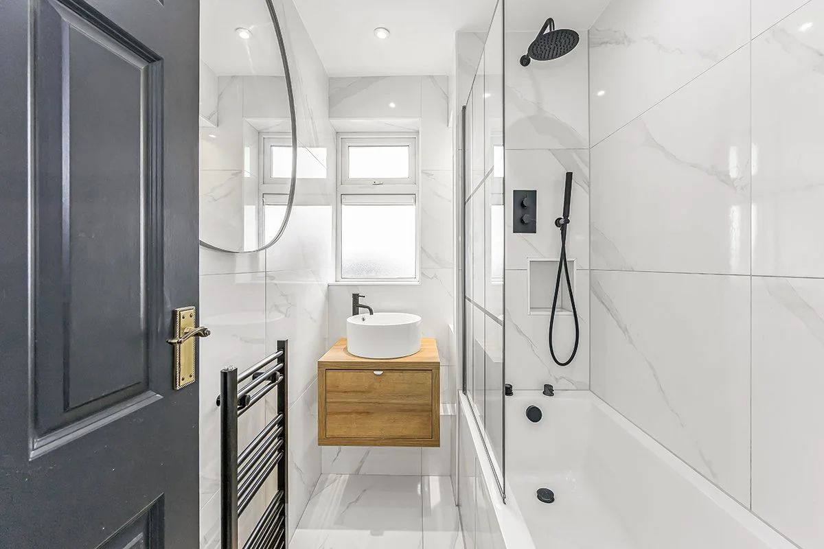 Modern small bathroom with a white bathtub, black shower fixtures, a floating wooden vanity with a white vessel sink, a large mirror, gray door, and a frosted window.