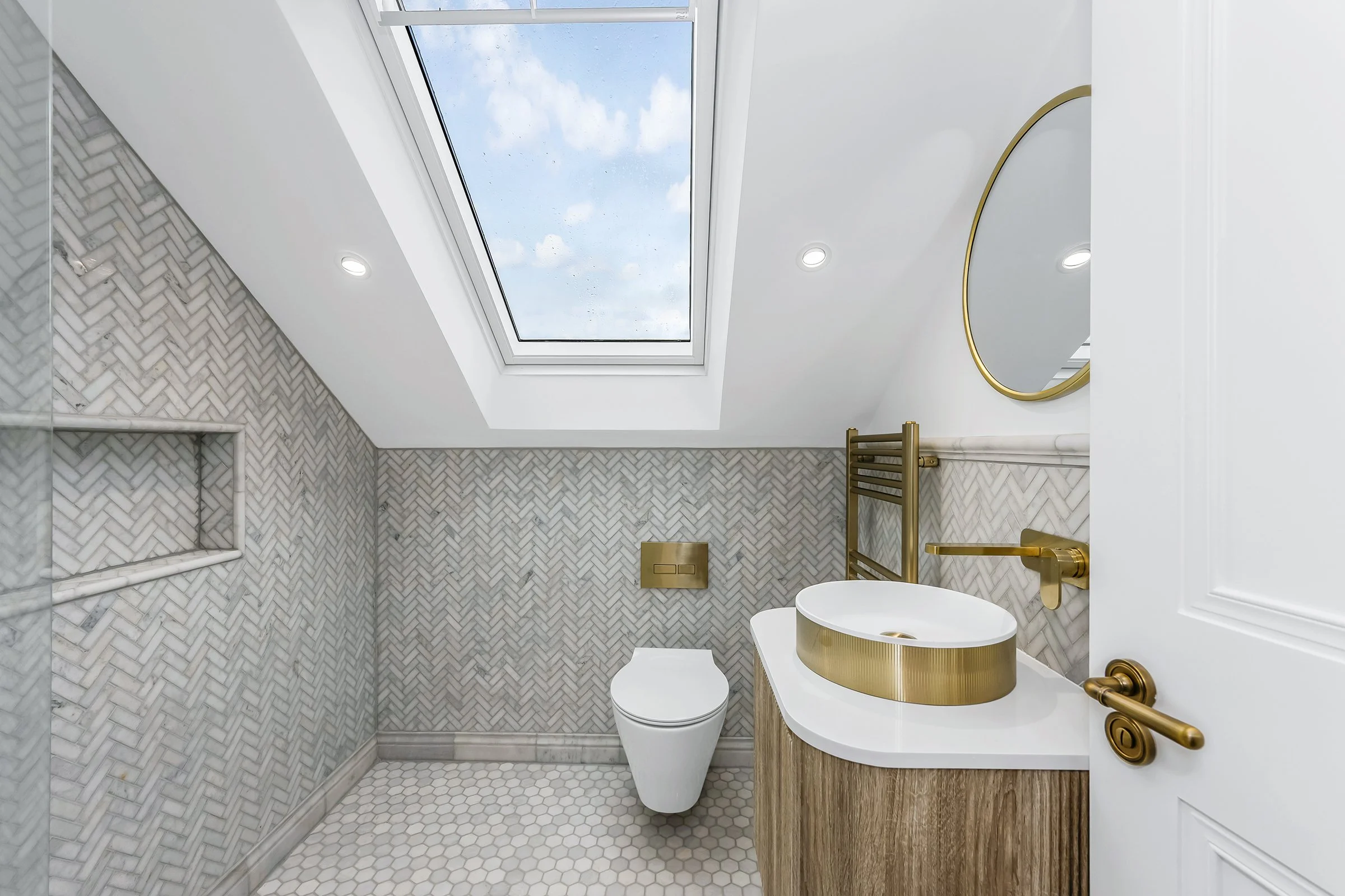A modern bathroom with a skylight showing a blue sky with clouds. The wall features gray herringbone tile, and there is a white toilet, a gold sink, a round gold mirror, and gold fixtures. The floor has small white hexagonal tiles.