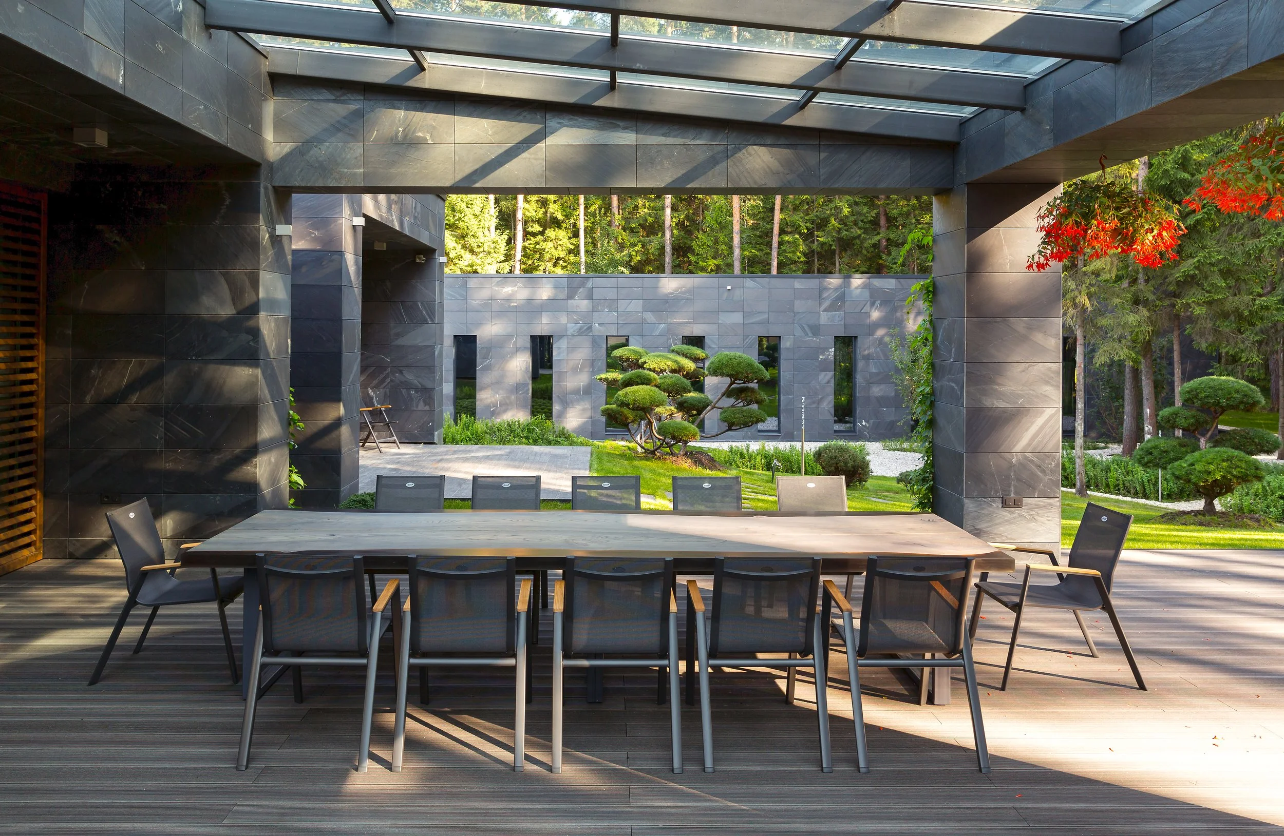 Modern outdoor patio area with a large dining table, black chairs, and a view of landscaped garden with trimmed bushes and trees in the background, beneath a partially covered deck with a glass roof.