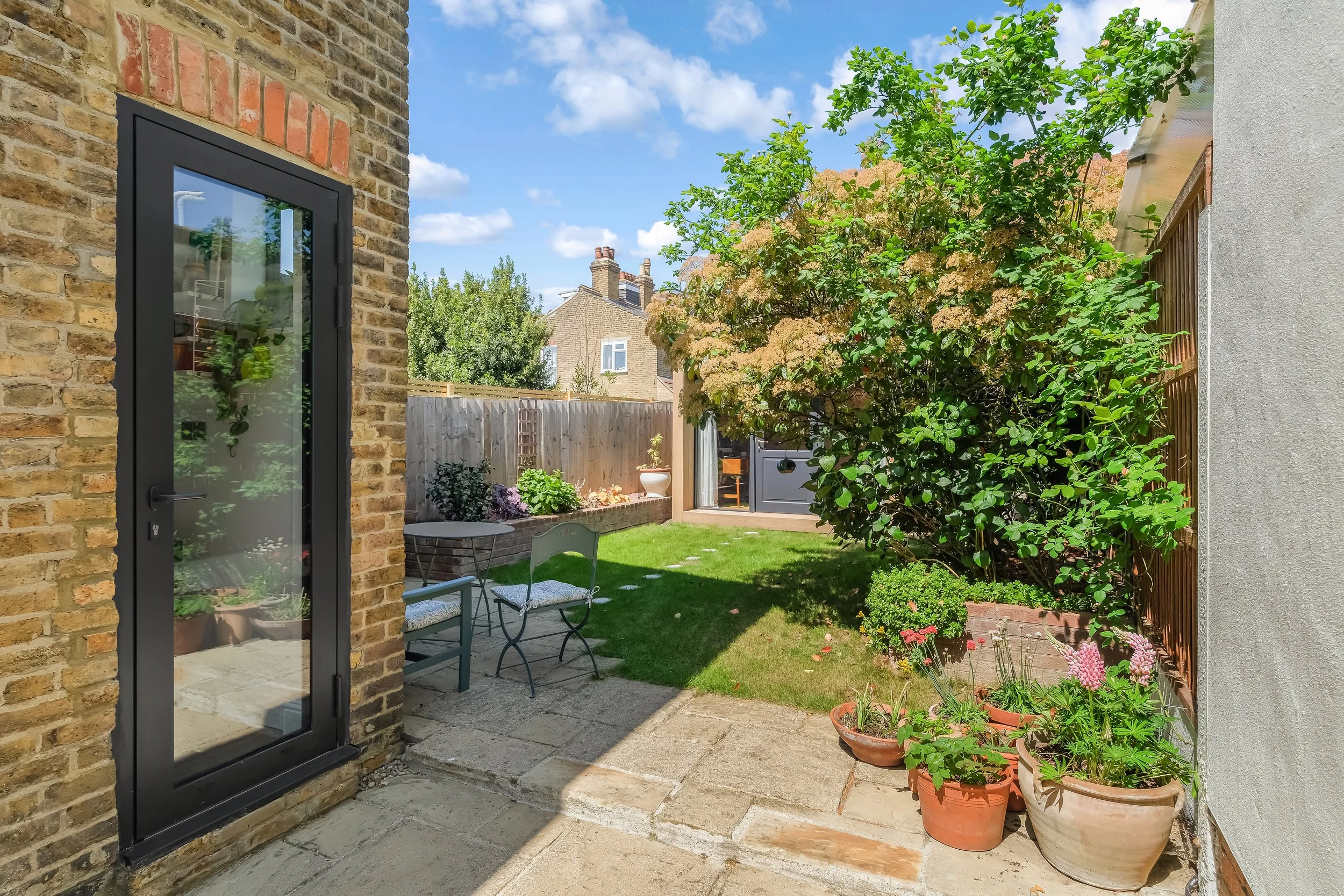 A small backyard with a brick house on the left, a garden with potted plants, a green lawn, and a large tree with green leaves. There is a gray shed and a patio area with a small table and chairs, under a blue sky with some clouds.
