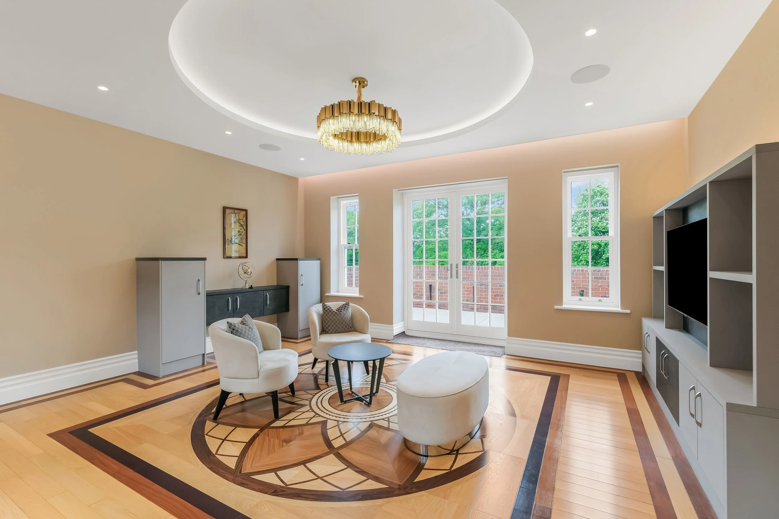 Interior of a modern living room with a wooden floor, beige walls, white ceiling, a large window, and a glass door. Features include a chandelier, white chairs, a black side table, and a gray shelving unit.