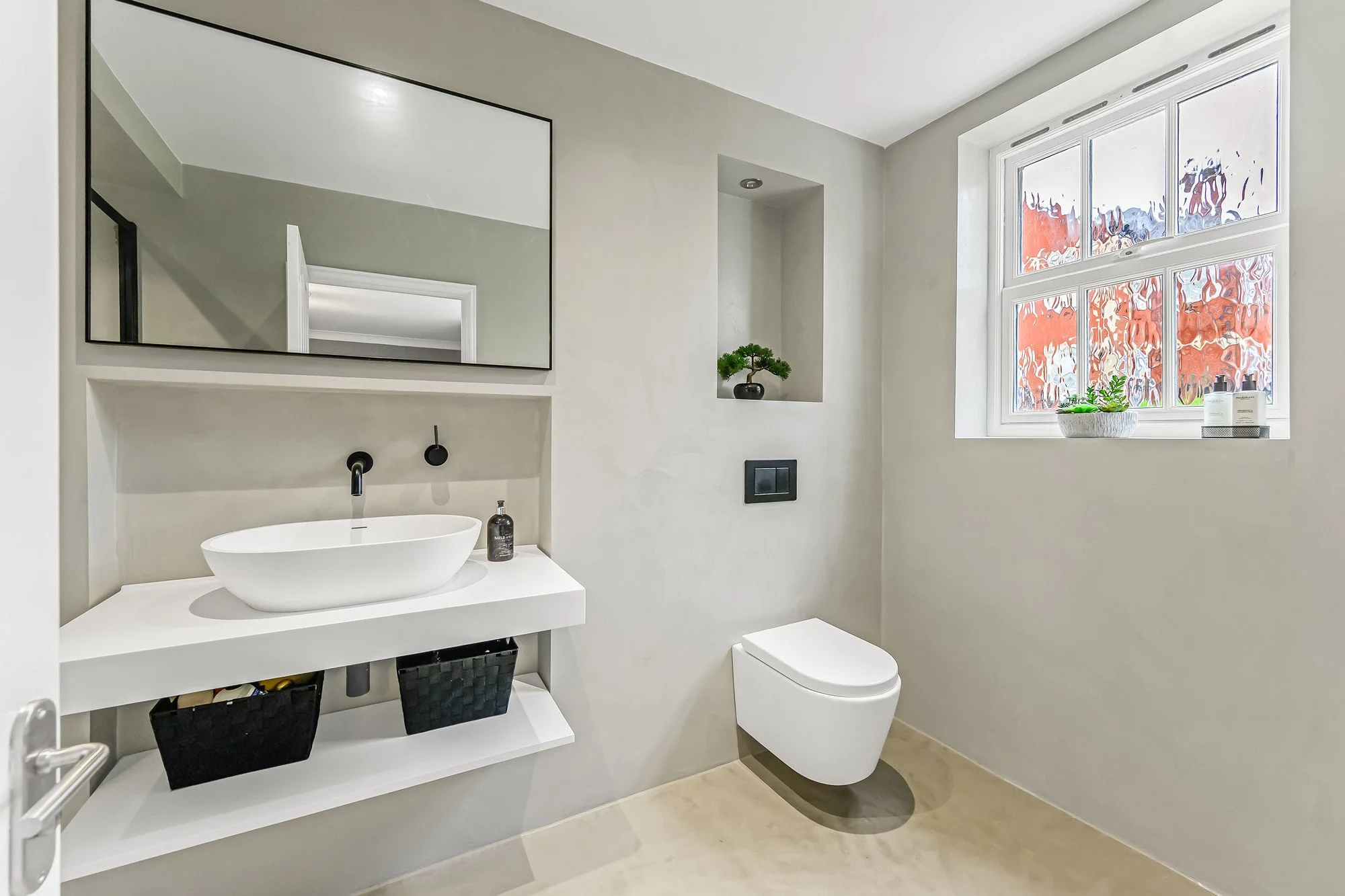 Modern bathroom with a white floating sink, black faucet, wall-mounted toilet, large mirror, and a window with a view of trees with red leaves. Decor includes a small bonsai and a soap dispenser.