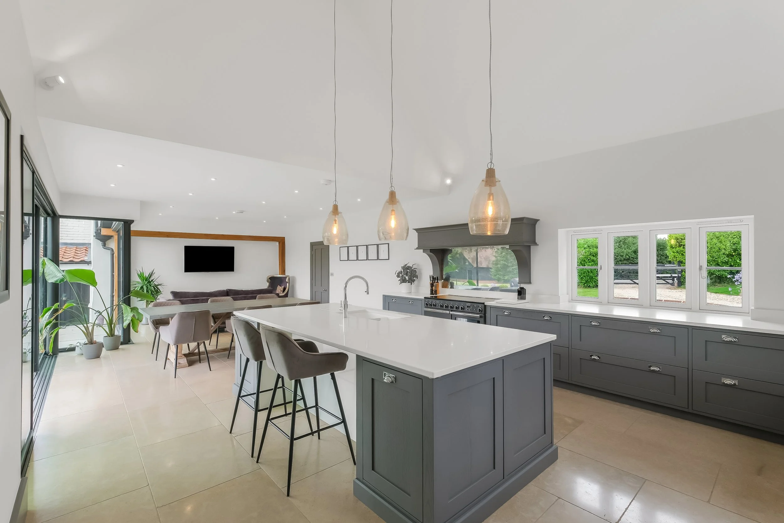 Bright, open-concept kitchen and dining area with gray cabinetry, white countertops, pendant lights, and large windows showing greenery outside.