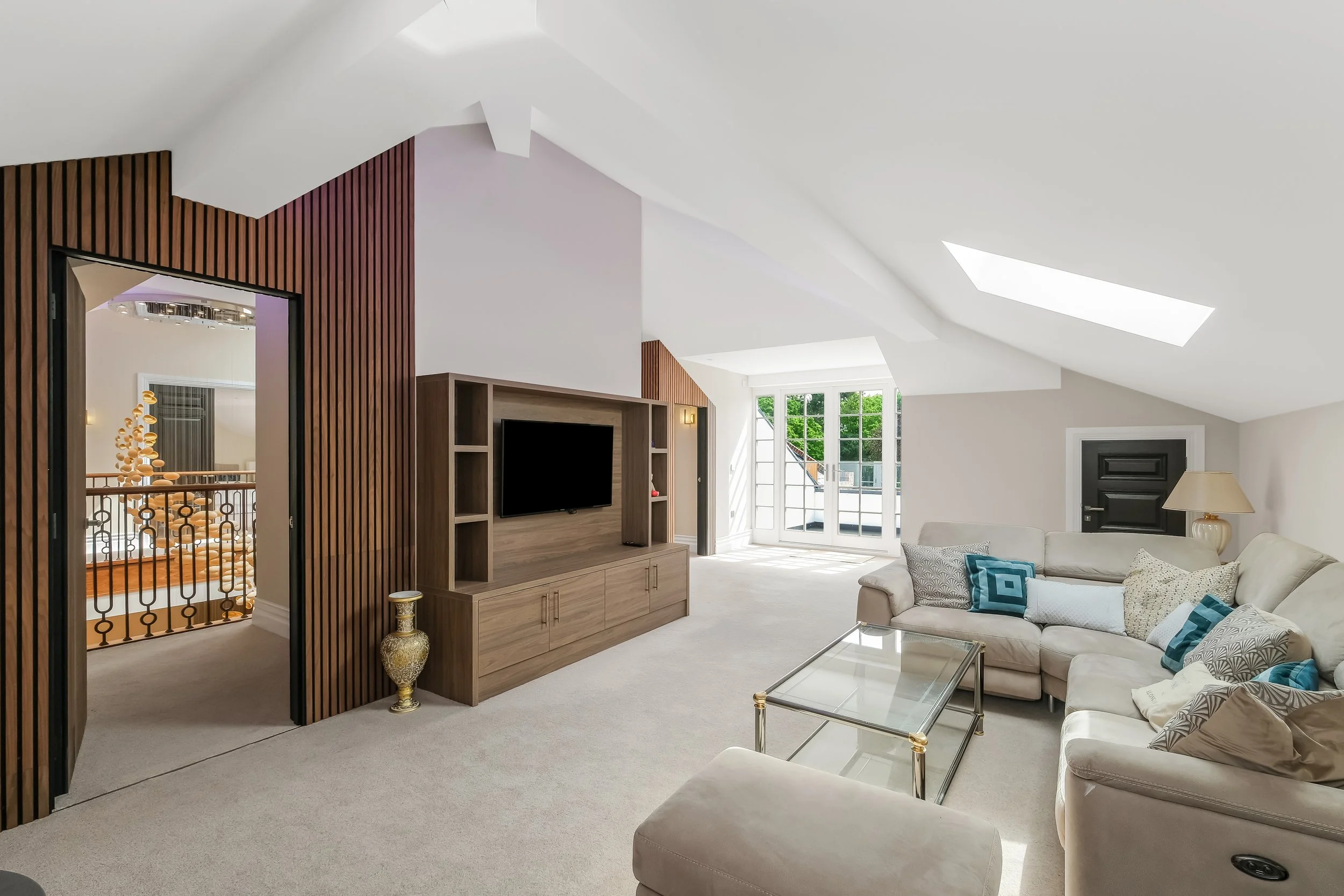 Bright living room with white walls, a high vaulted ceiling with skylights, a beige sofa with blue and patterned pillows, a wooden media console with a television, large windows, and a doorway leading to a staircase.