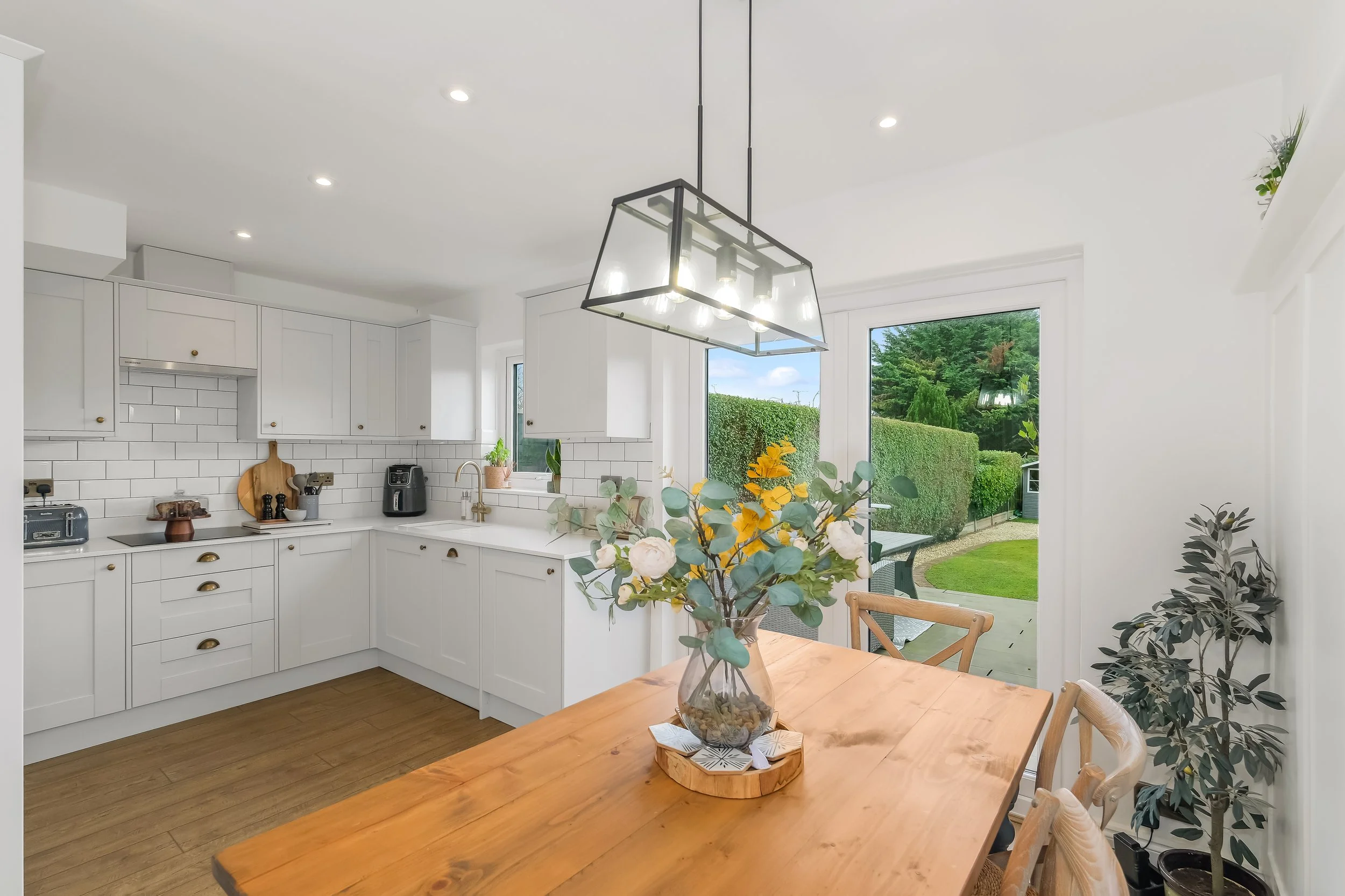 A bright kitchen with white cabinets and a wooden table with a flower arrangement. Large windows reveal a green backyard with trees and a trimmed hedge. Recessed ceiling lights and a modern rectangular pendant light hang over the space.