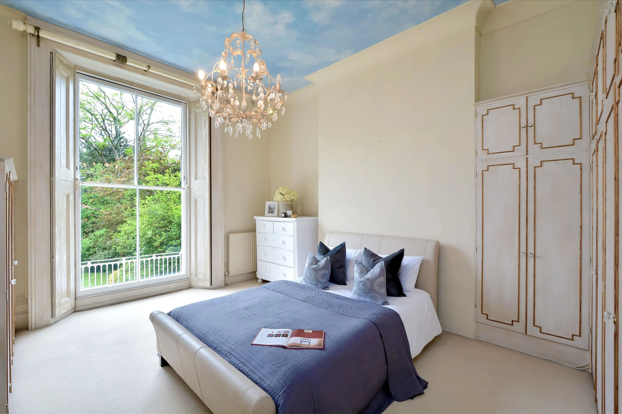 Bright bedroom with large window showing greenery outside, white bed with gray bedding and pillows, white dresser, floral decor, and chandelier ceiling light, with painted sky ceiling.