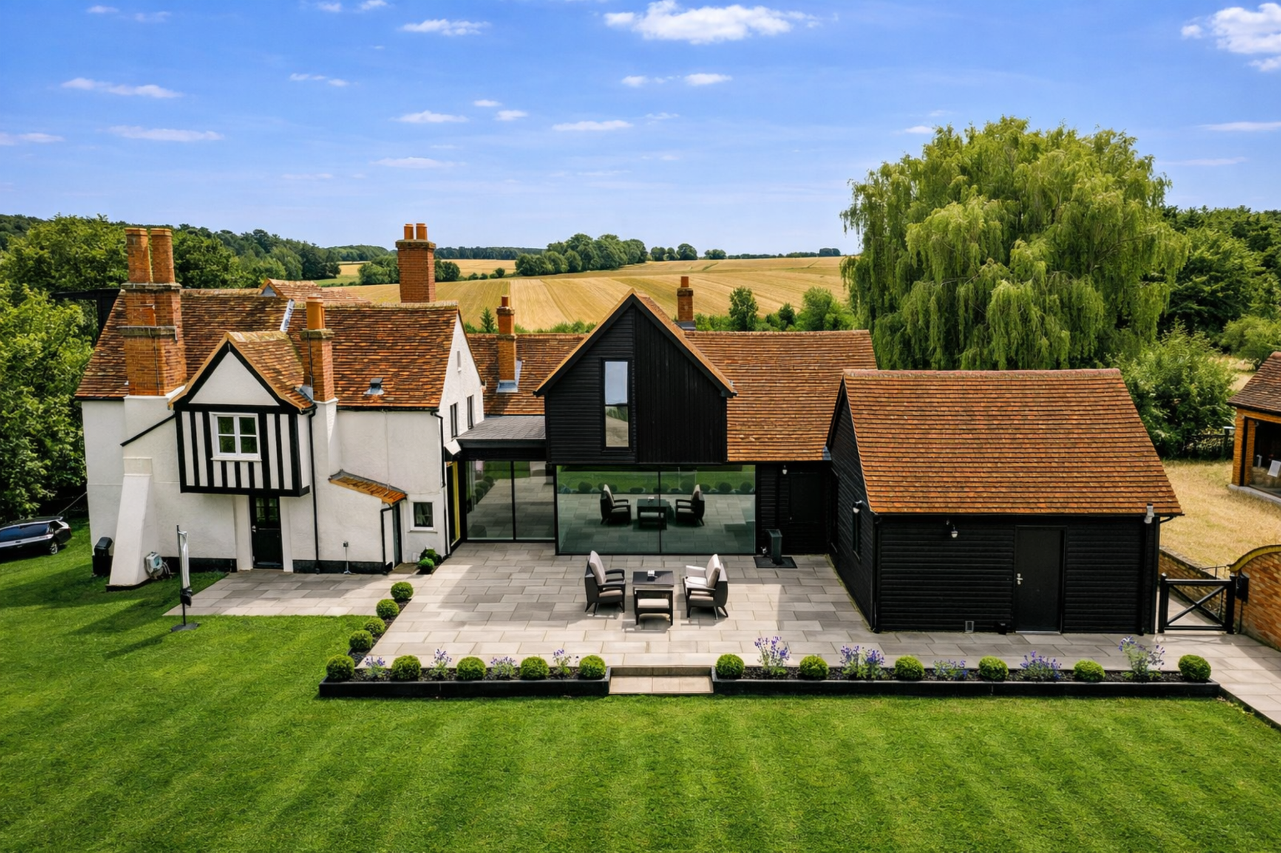Modern house with black and white exterior, large glass patio, outdoor dining set, and well-manicured lawn with flower bed, set in a rural landscape with fields and trees under a partly cloudy sky.