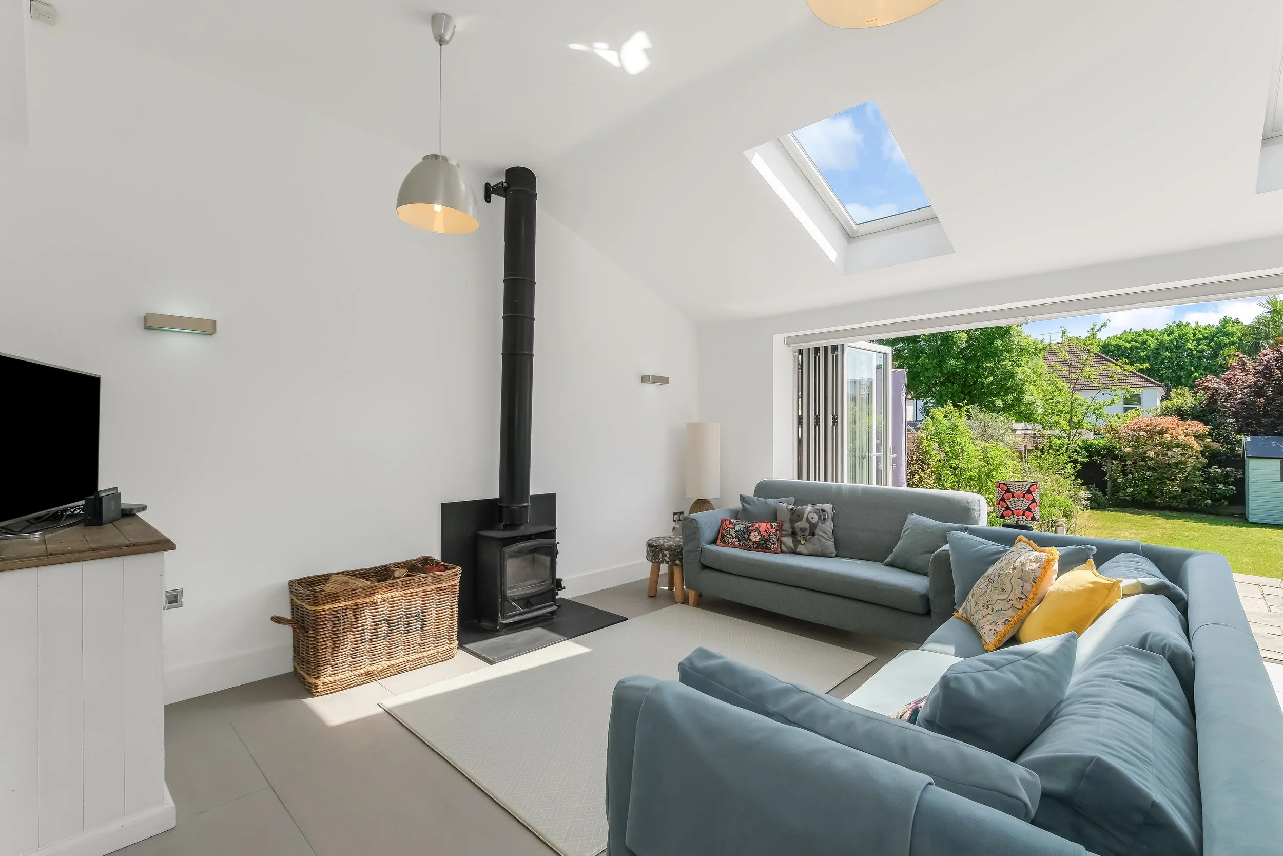 A bright living room with a pale blue sofa, colorful cushions, a wood-burning stove, a skylight showing a blue sky with clouds, a large window showing green trees and grass outside, and a small side table with a lamp.