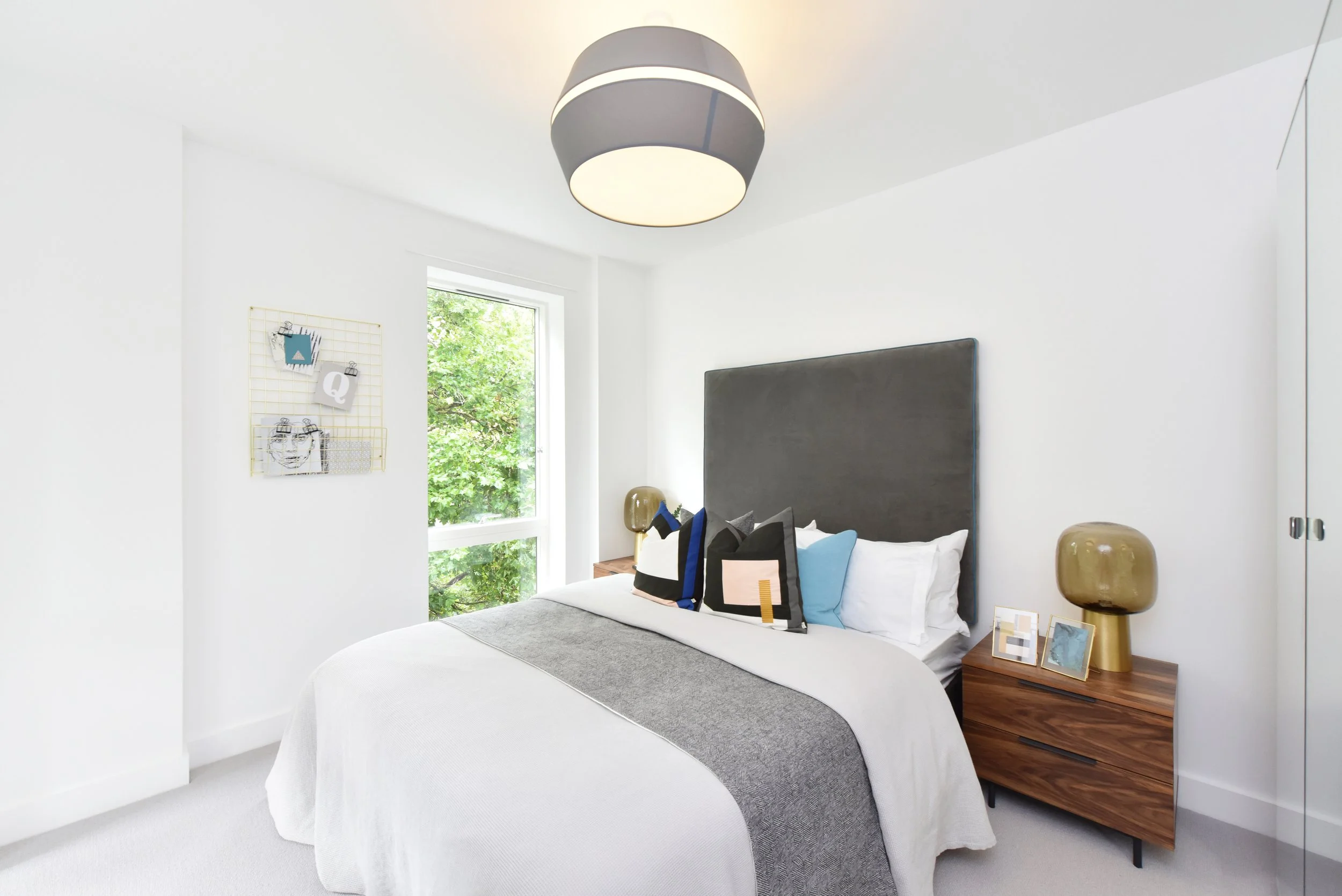A modern bedroom with a bed featuring white linens, a gray blanket, and decorative pillows in black, white, and blue. There are two nightstands with gold lamps, framed pictures, and a wooden dresser. A window shows green trees outside, and a ceiling 
