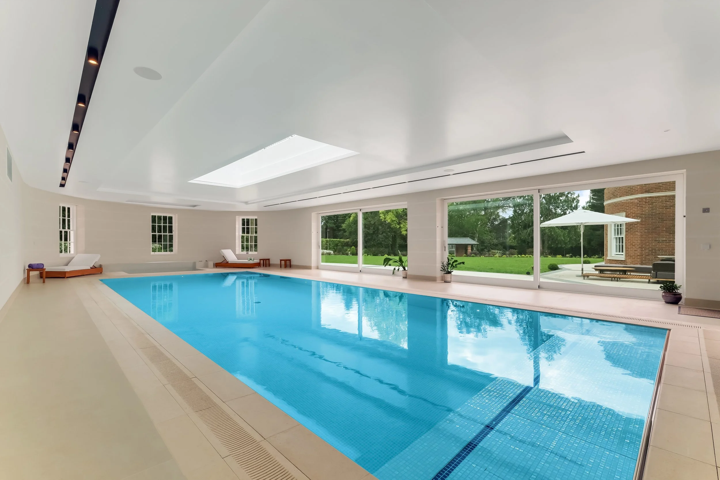 Indoor swimming pool area with large glass windows, white walls, and a view of a green backyard with trees and a patio umbrella.