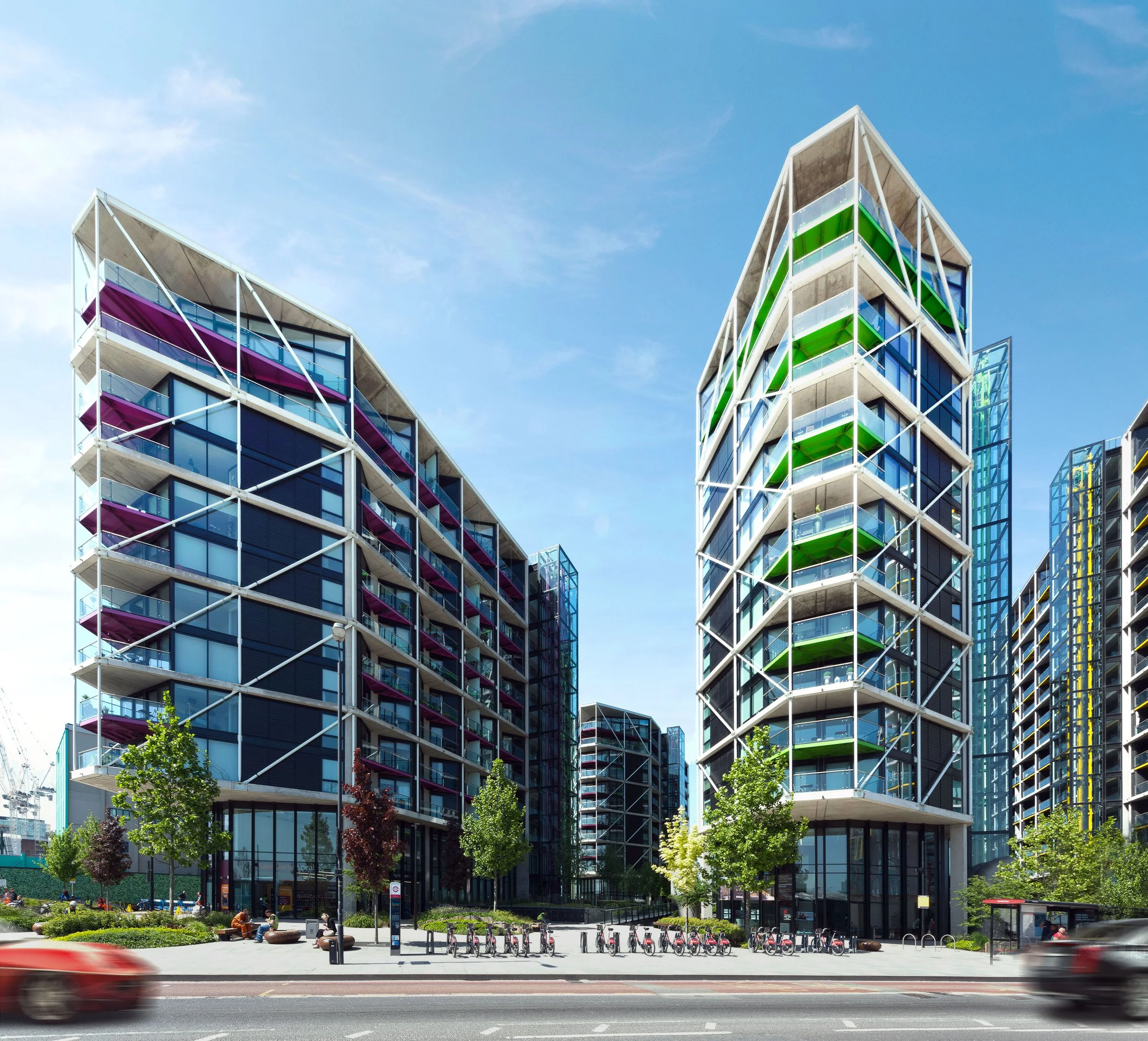 Modern high-rise buildings with colorful balconies, surrounded by green trees, people sitting on benches in a landscaped park area, and bicycles parked near the sidewalk, with a vehicle in motion on the street in the foreground.