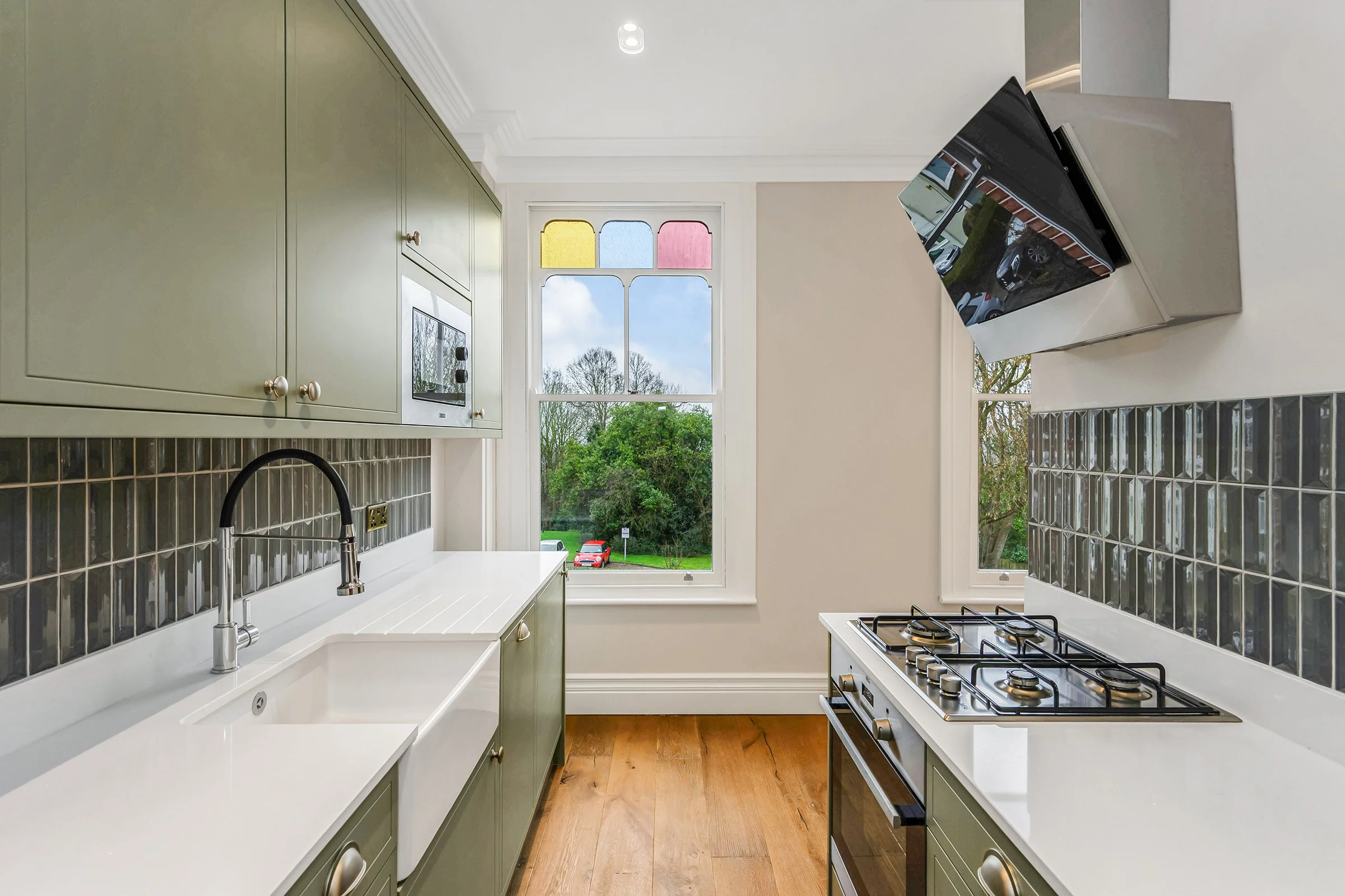 A bright kitchen with green cabinets, a white countertop, a window with colorful stained glass at the top, and a view of trees and parked cars outside.