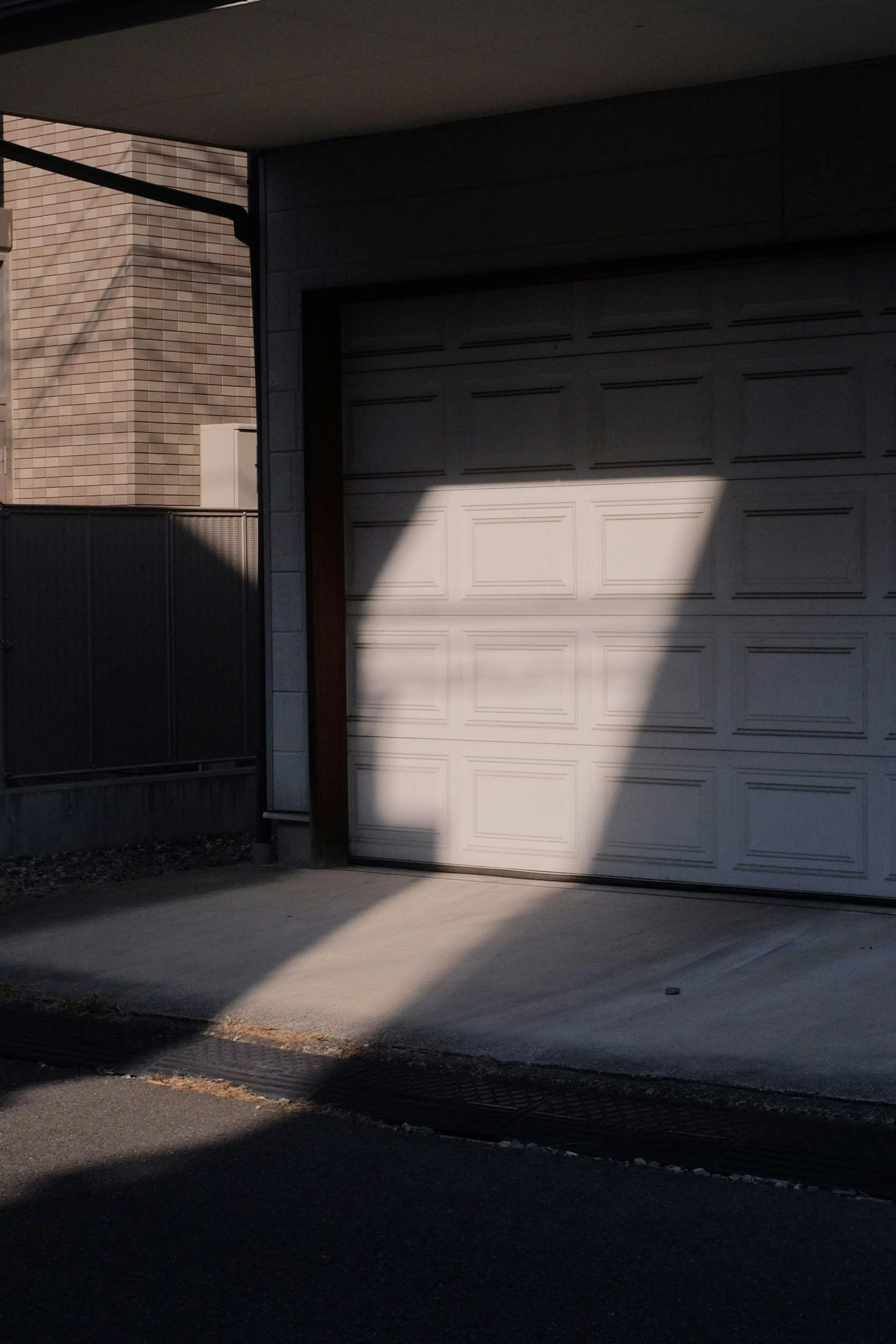 A closed garage door casting a shadow on the driveway and concrete floor in front of it, with part of a brick wall and fence visible on the left side.