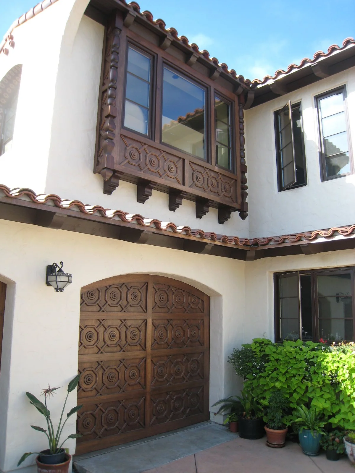 A two-story house with white stucco walls, a red tile roof, and wooden window frames and shutters. There is a wooden garage door with decorative carvings, a black outdoor wall lantern, and potted plants near the entrance.