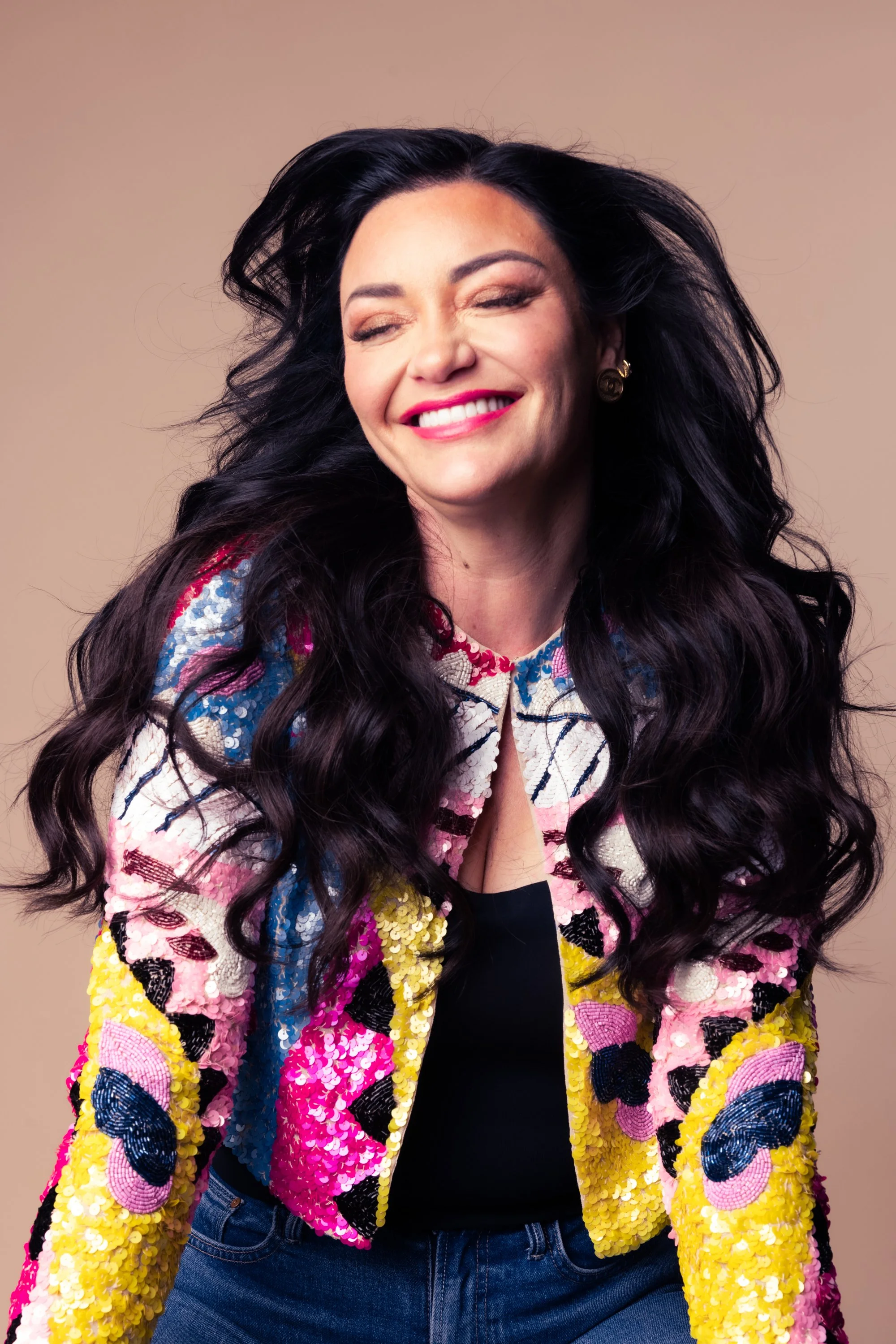A woman with long dark wavy hair, smiling with eyes closed, wearing a colorful sequined jacket and blue jeans, against a beige background.