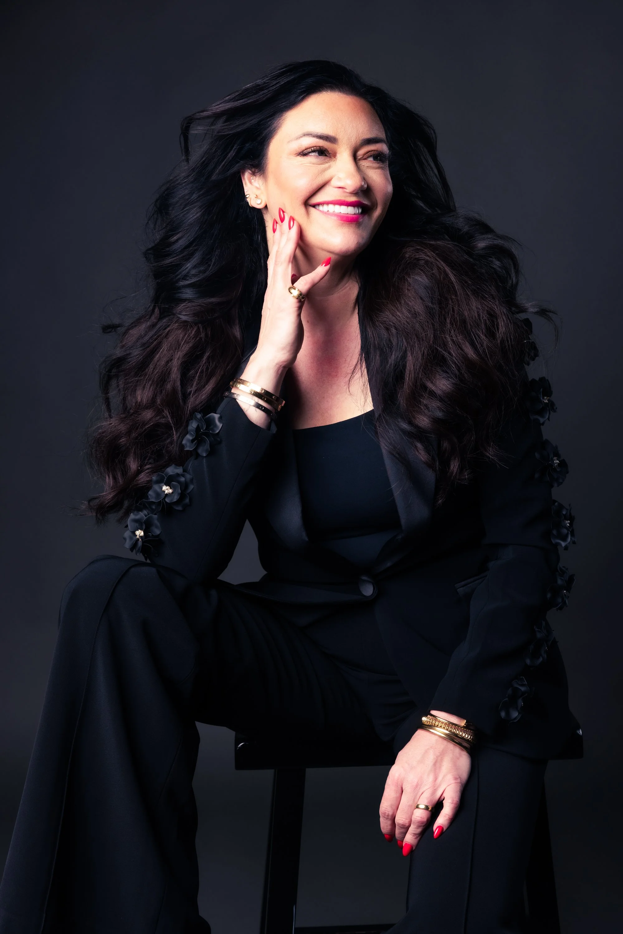 A woman with long dark wavy hair, wearing a black outfit with floral embellishments on the sleeves, sitting against a dark background, with red nail polish, gold jewelry, and smiling while touching her face.
