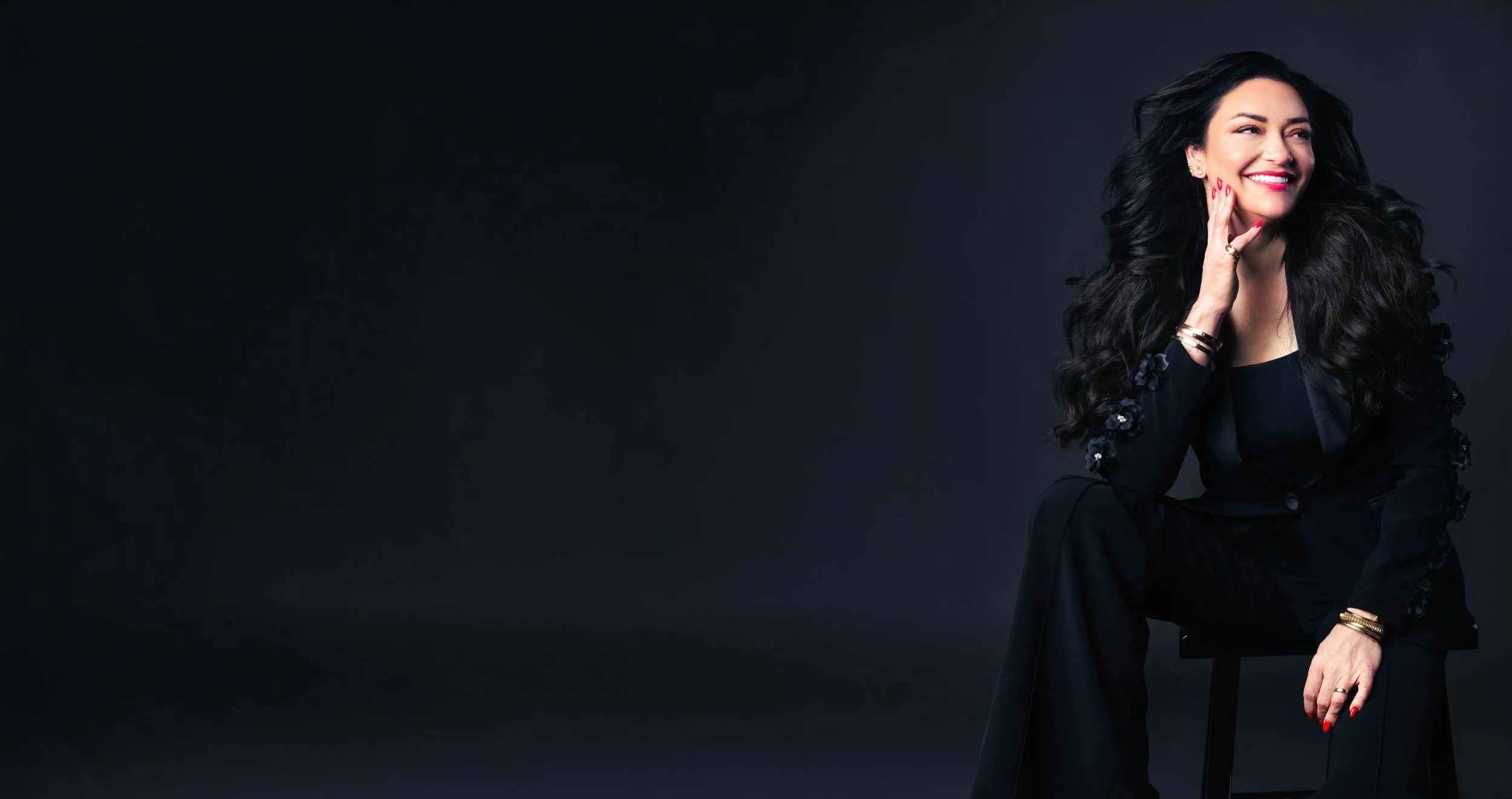 A woman with long black wavy hair sitting on a black chair against a dark background, smiling and looking to her right.