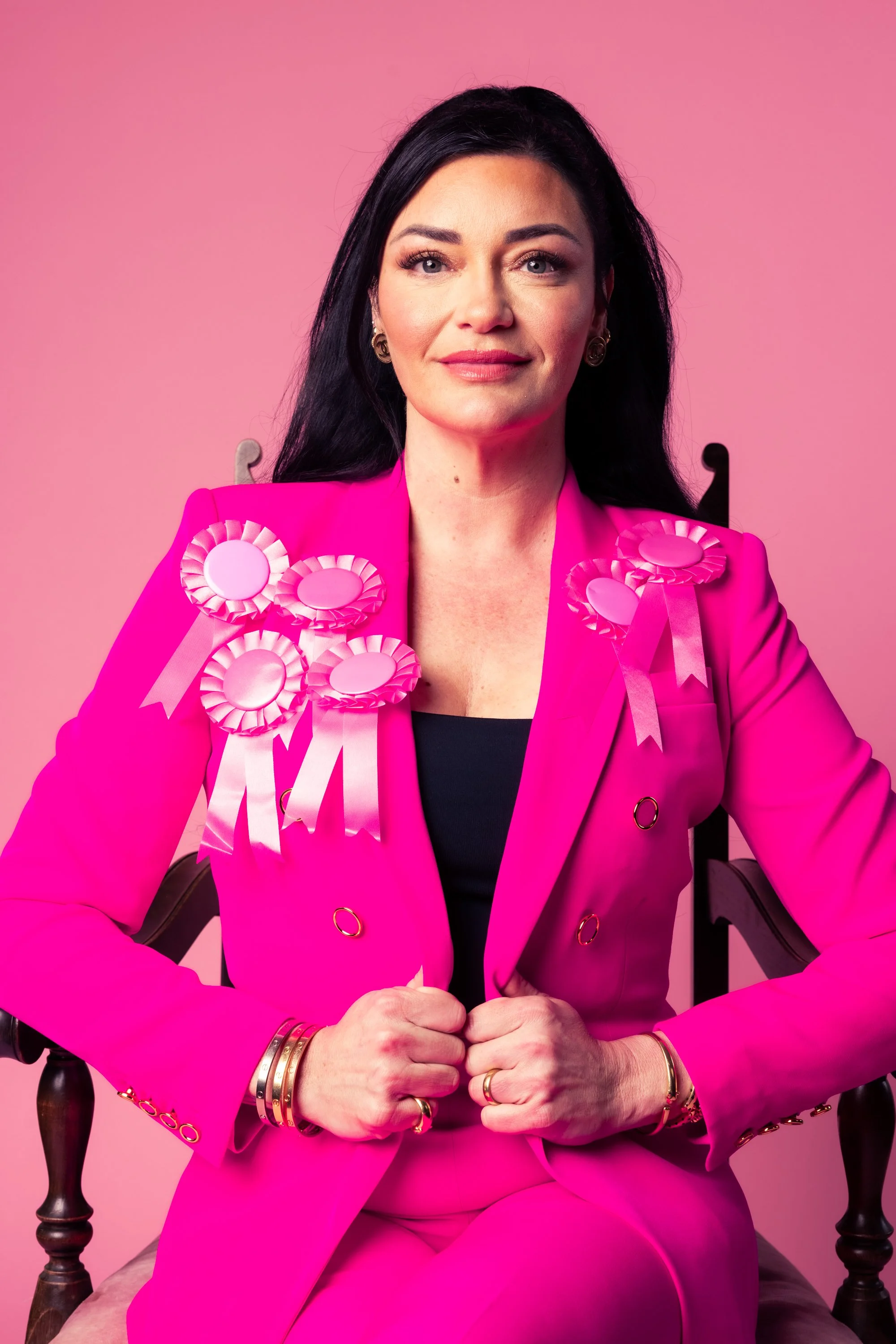 A woman with long black hair, wearing a bright pink suit jacket adorned with pink ribbons and rosettes, sitting in a black wooden chair against a pink background.