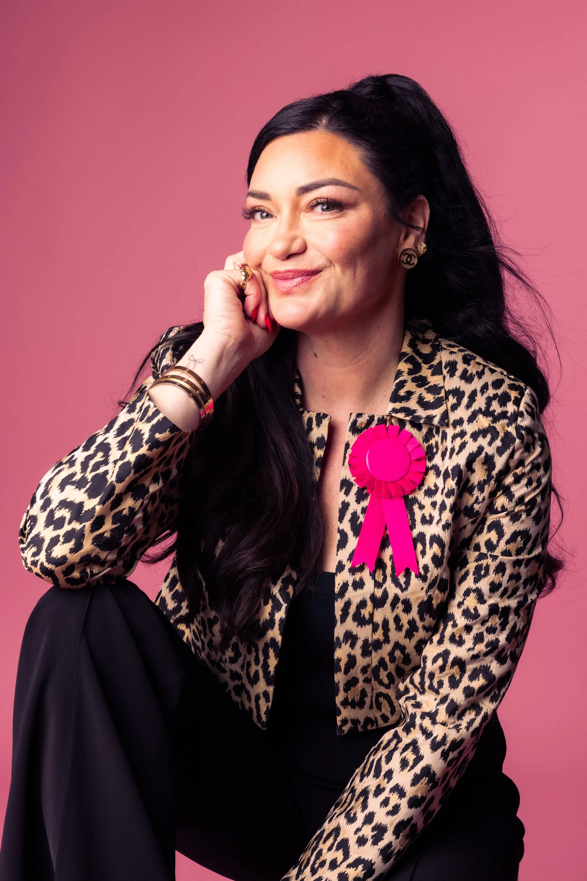 A woman with black hair, gold jewelry, and a leopard print blazer winks at the camera against a pink background. She has a pink ribbon pinned to her blazer.