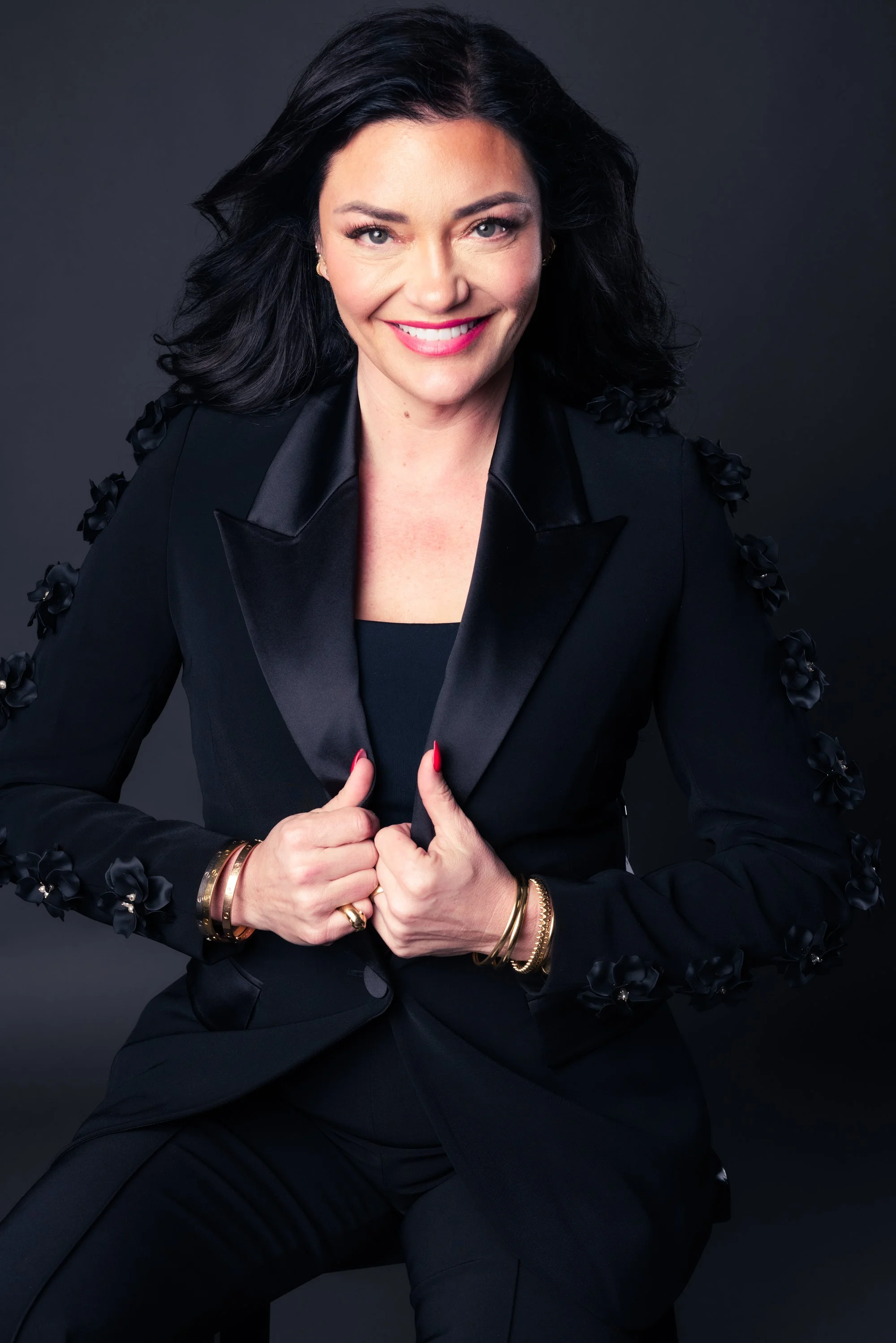 A woman with black hair, wearing a black suit with floral embellishments on the shoulders, smiling and holding the lapels of her blazer with both hands, against a dark background.