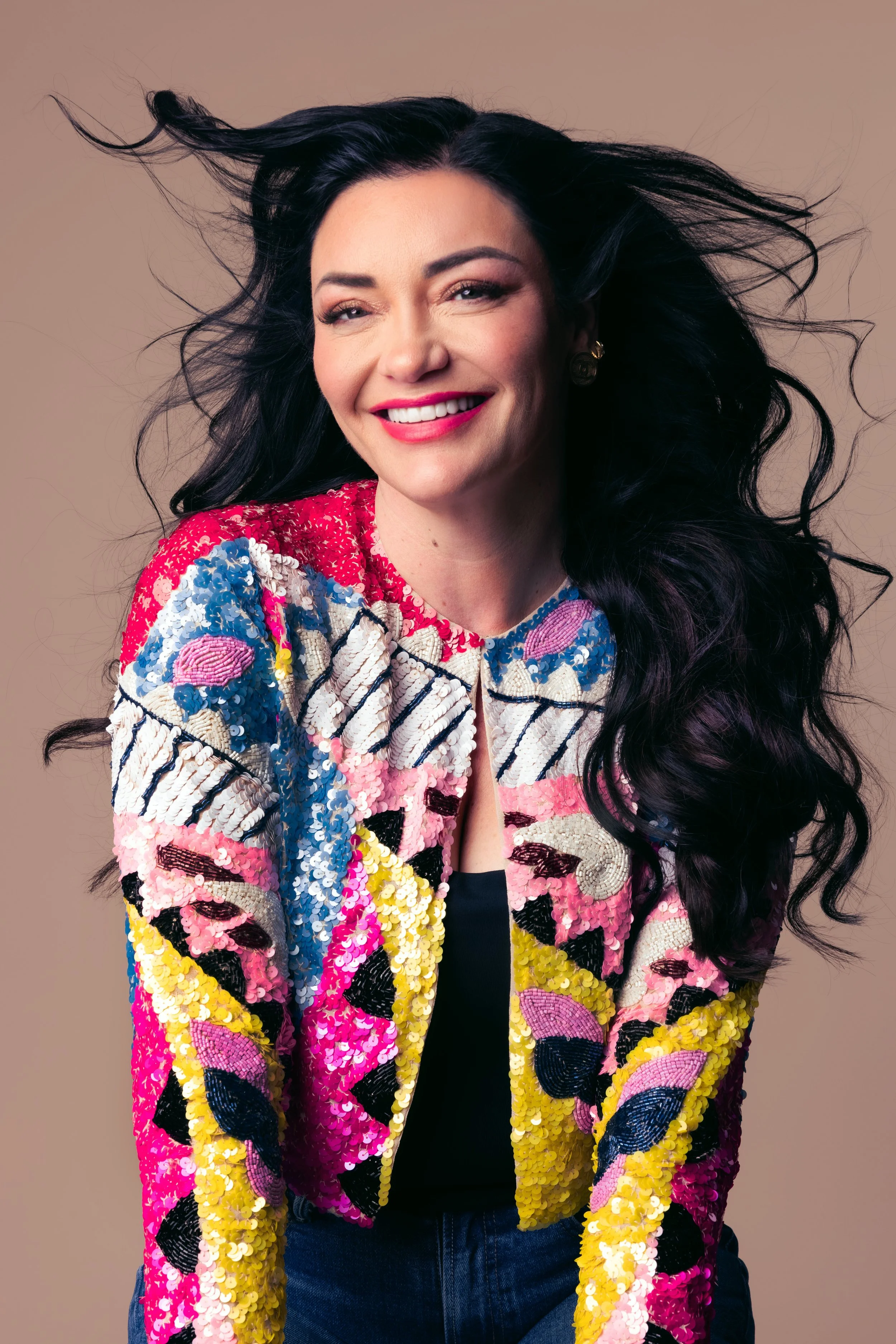 A woman with long dark hair smiling, wearing a colorful sequined jacket with pink, yellow, black, and white patterns, and a black top underneath, against a neutral background.