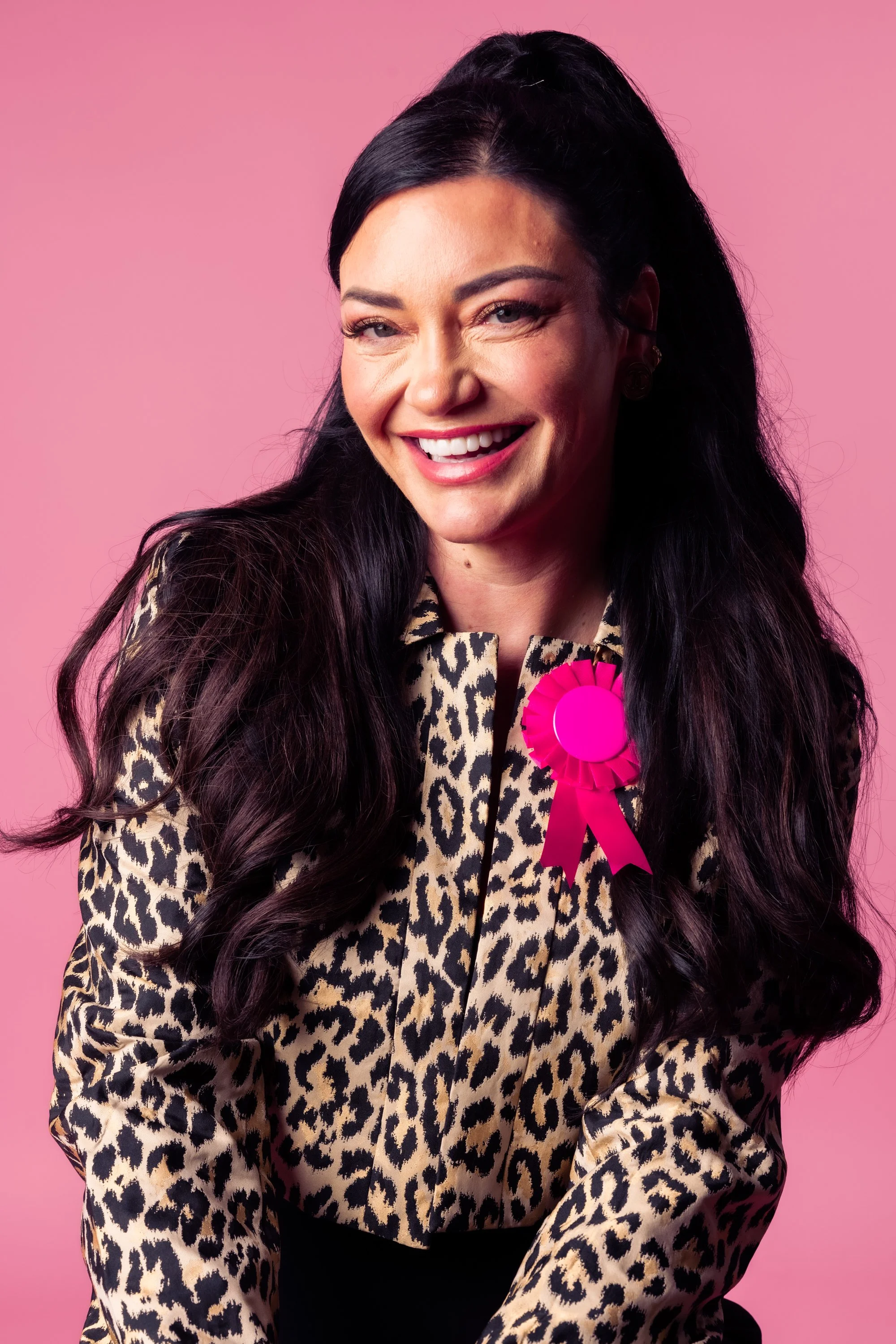 A woman with long dark hair, wearing a leopard print shirt, smiling, with a pink ribbon on her shirt, standing against a pink background.