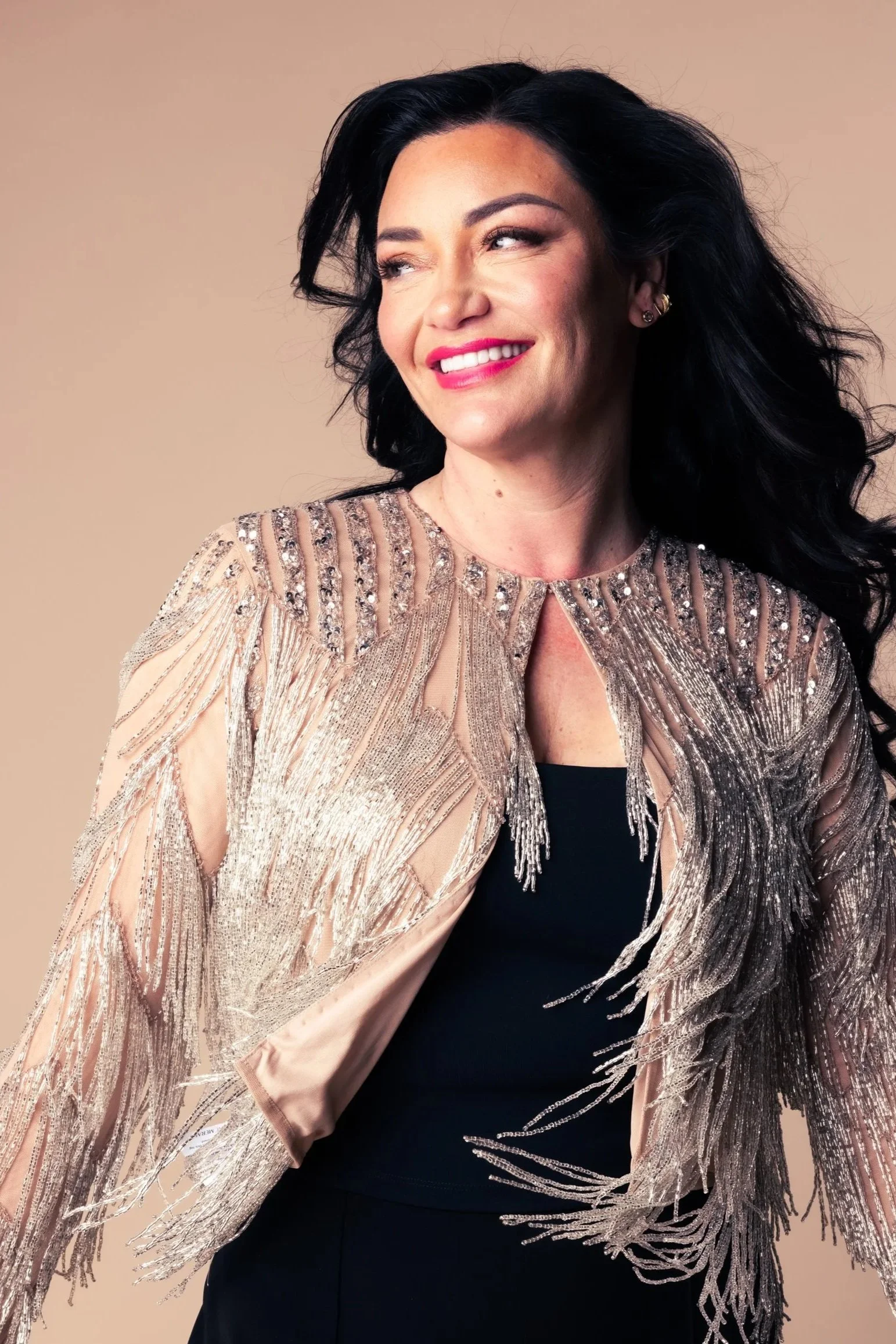 A woman with dark, wavy hair, wearing makeup, and dressed in a black top with a glittering, beige, and silver embroidered jacket that has fringes.
