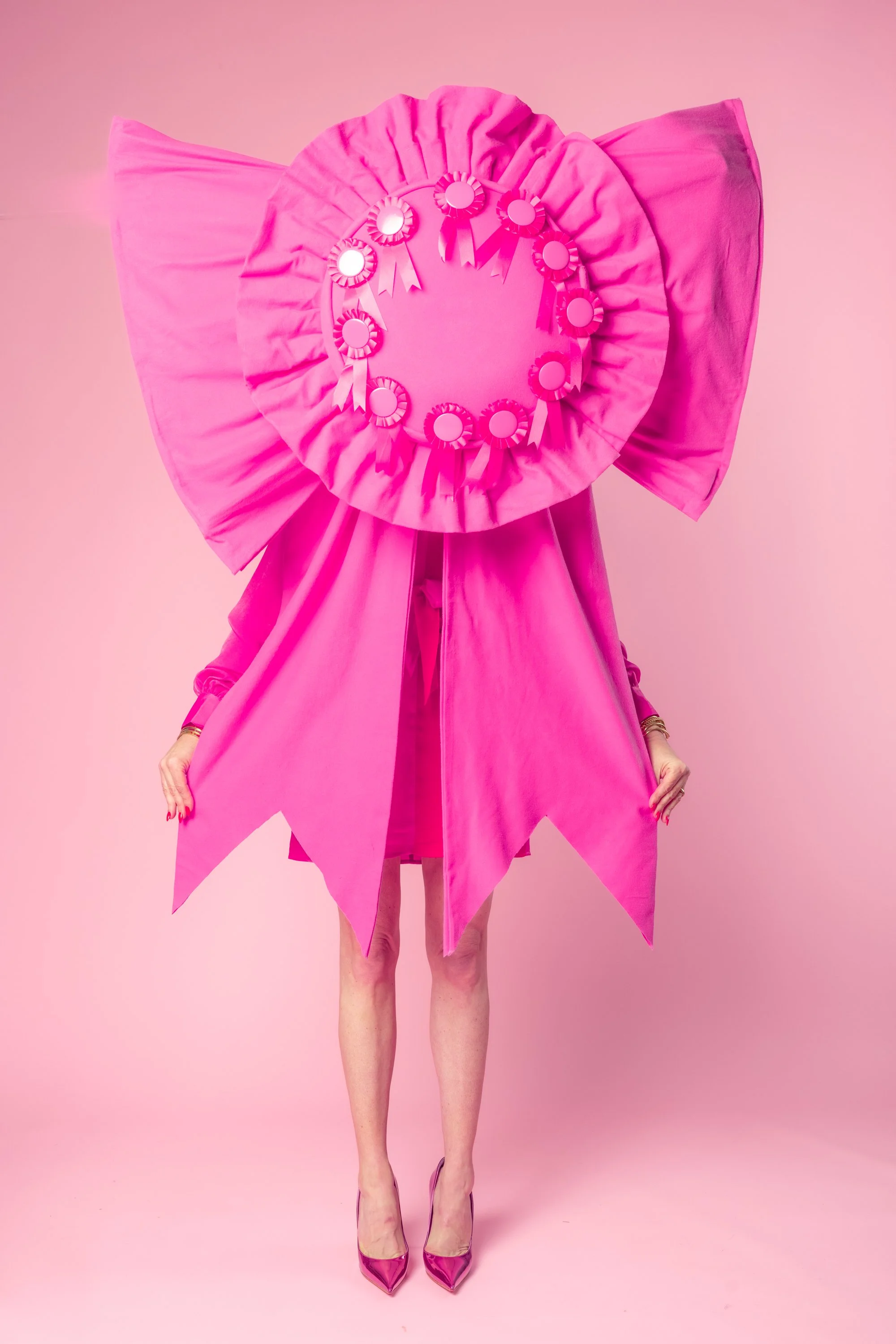 A person dressed in pink wearing a large pink bow with ribbons and rosettes, pink high heels, and standing against a pink background.