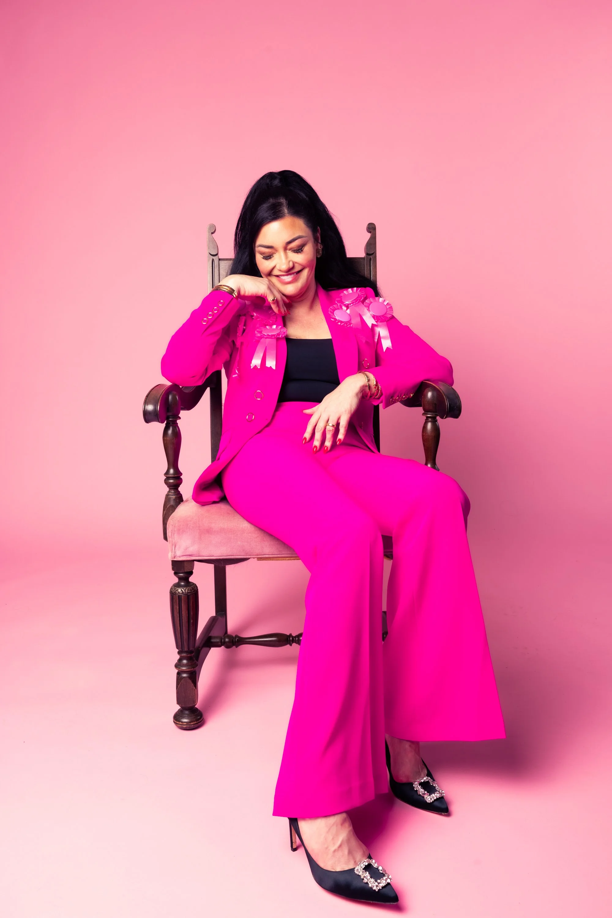 A woman with long dark hair sitting on a wooden chair against a pink background. She is wearing a bright pink suit with matching pants, a black top, and black high heels with sparkling embellishments. She is smiling and looking down with her eyes closed, with her hand resting on her face and her other hand on her lap. Pink ribbons with rosettes are attached to her suit lapels.