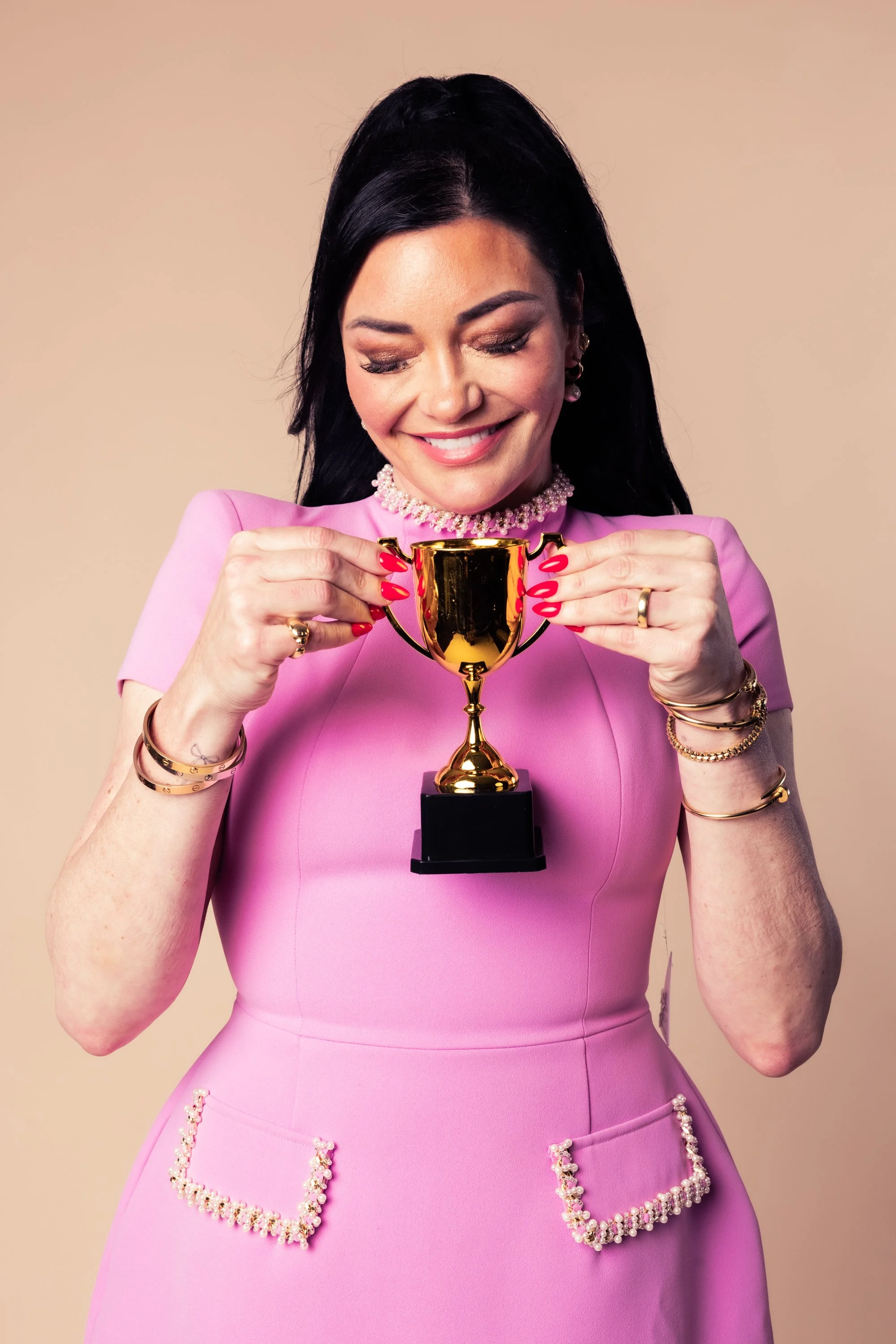 Woman in pink dress holding a gold trophy, smiling with eyes closed, with pearl jewelry and matching pink and pearl embellishments on the dress.