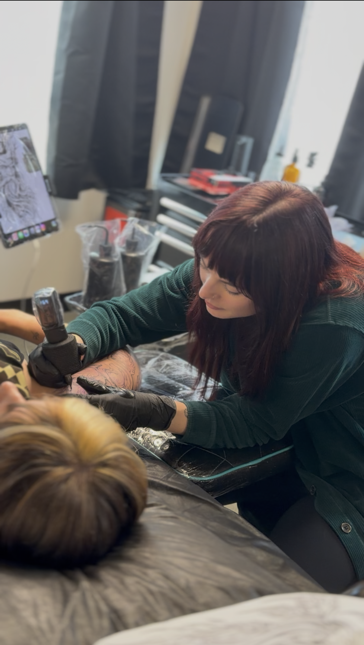 A woman with dark red hair in a tattoo studio doing a tattoo on a person's arm.