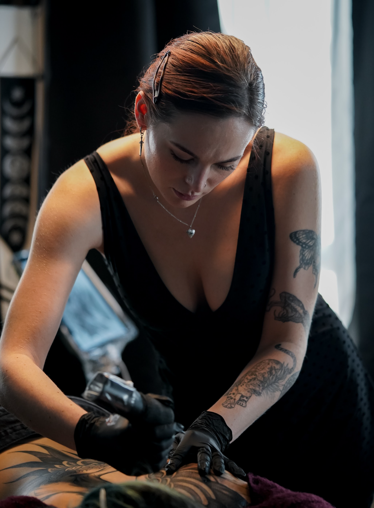 A woman with tattoos, earrings, and a heart necklace tattoos on her arm and chest, wearing a black top, receiving a tattoo on her thigh. She has glasses on her head and is focused on the tattooing process.