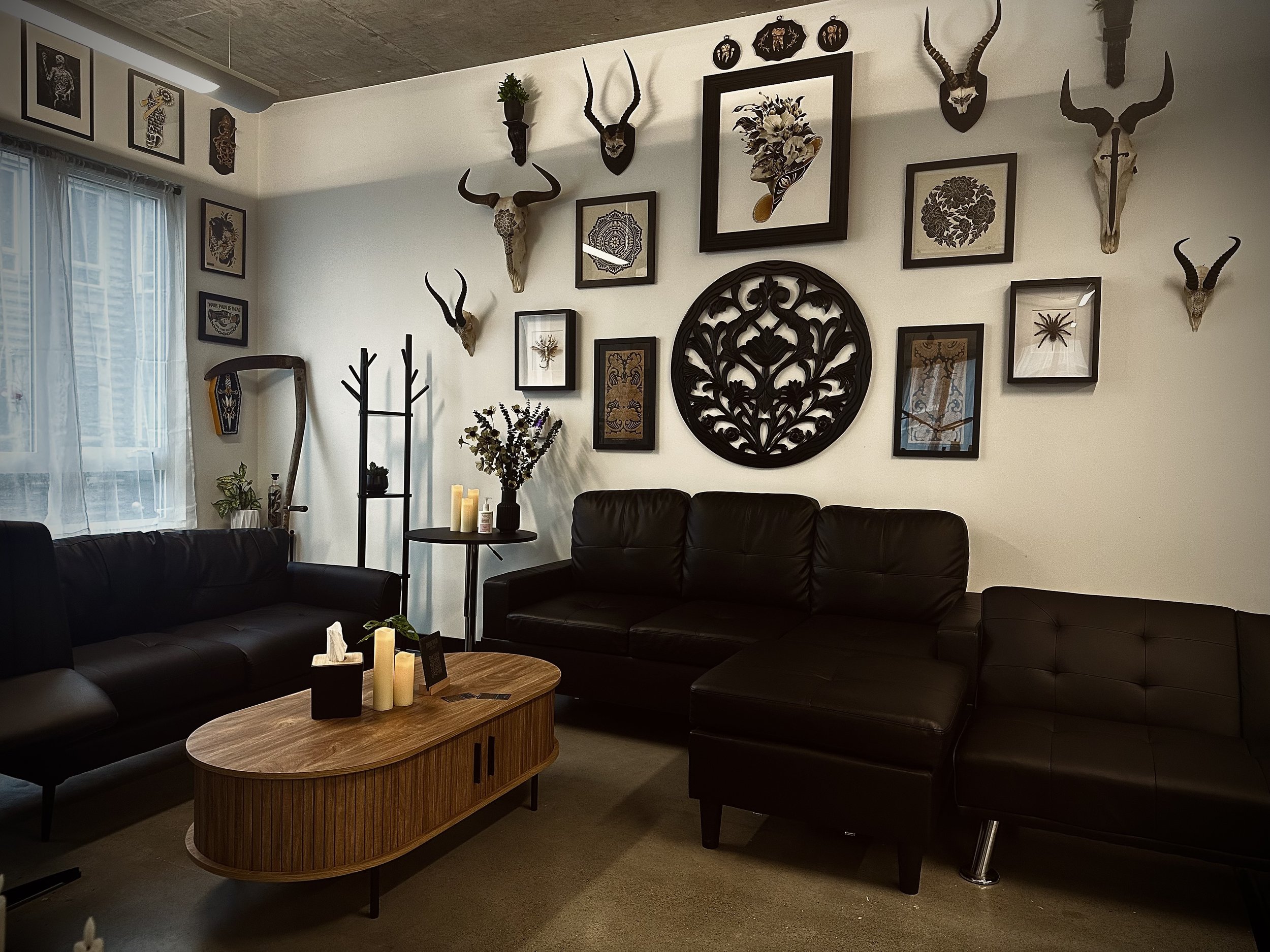 Living room with white wall decorated with multiple animal skulls and framed artwork, large window with white curtains, black leather sofas, wooden coffee table with candles and small decorative items, some plants and a coat rack.