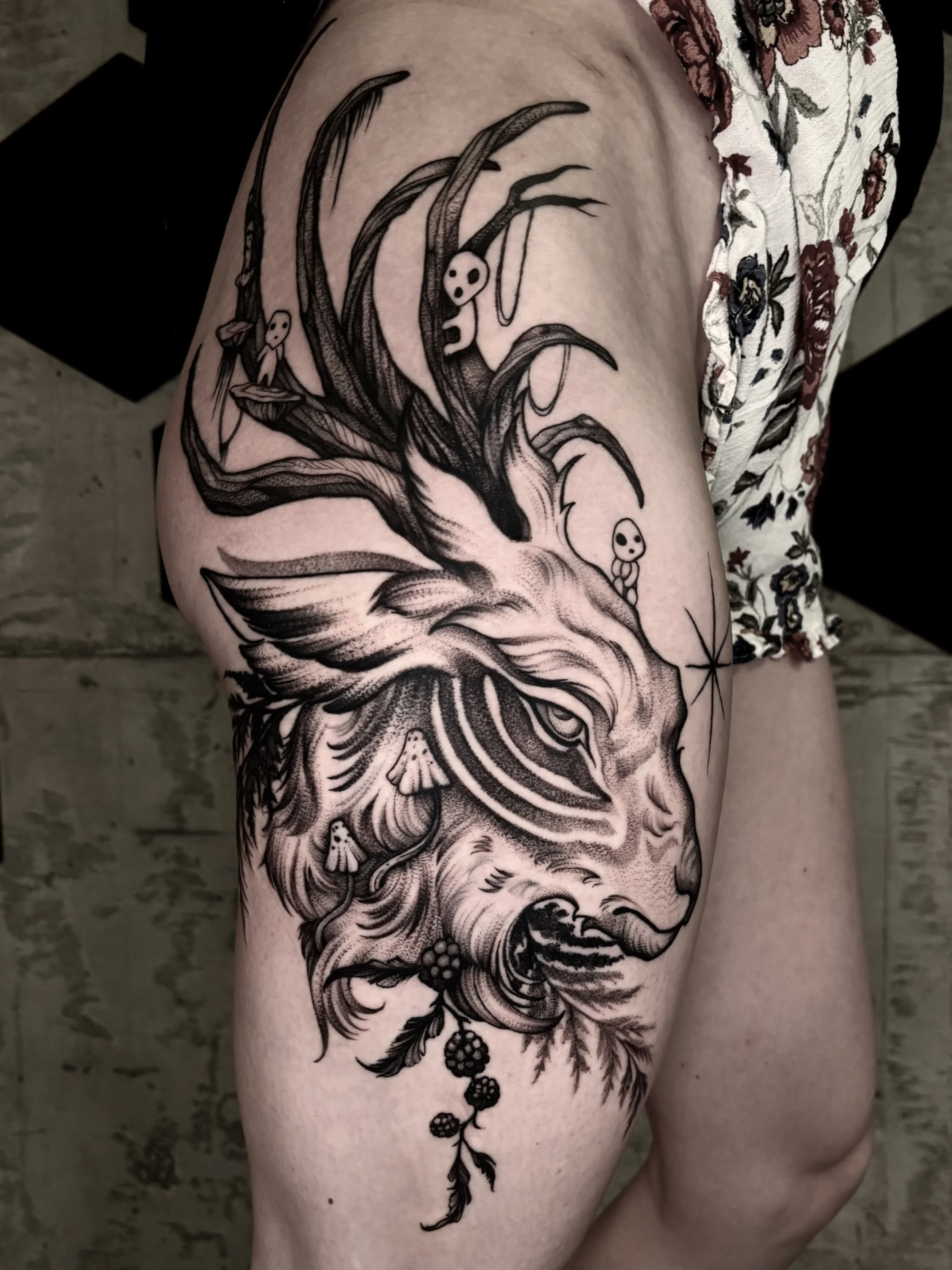 Black tattoo on person's thigh featuring a detailed wolf head, surrounded by floral and nature-inspired elements.
