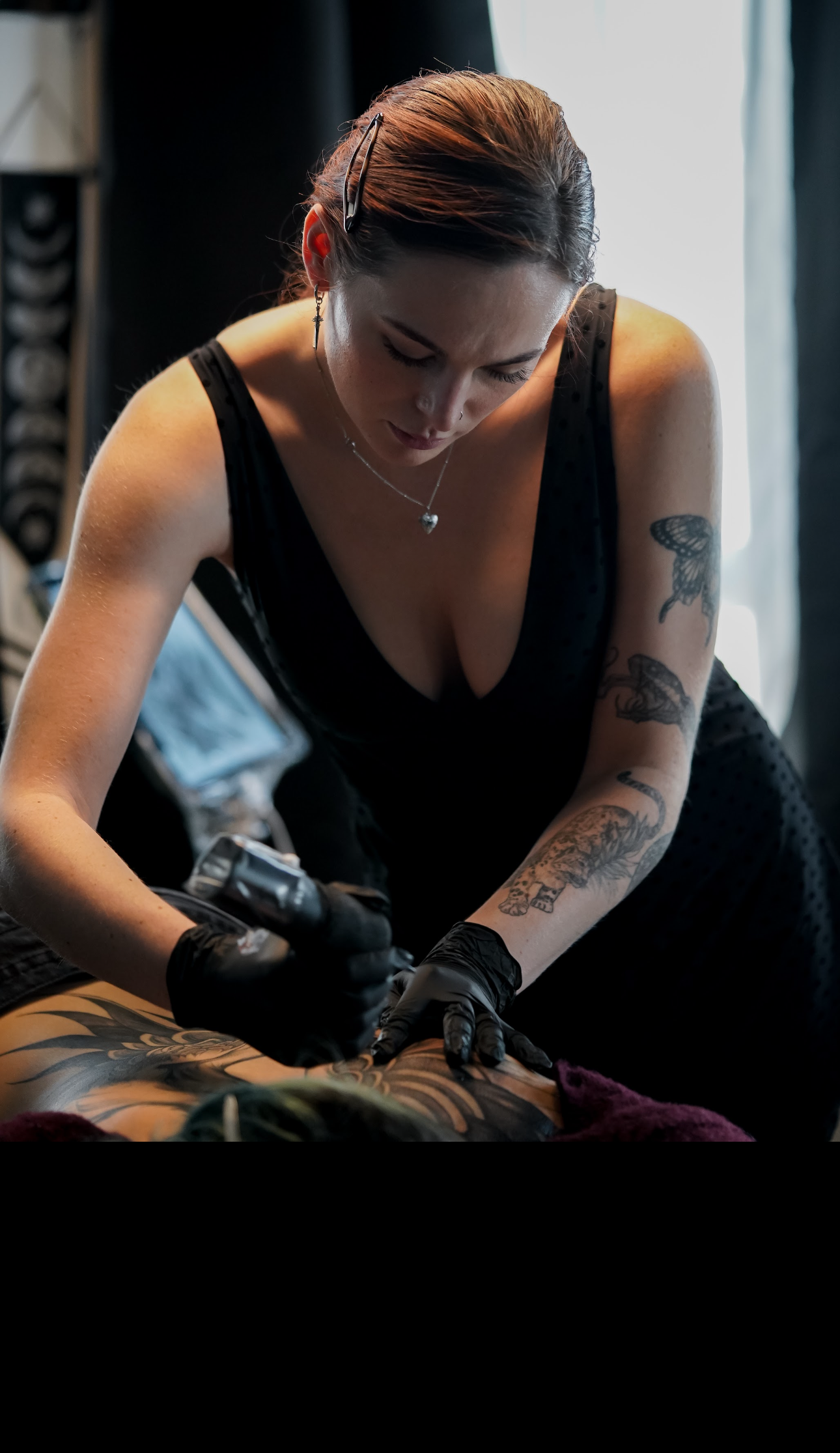 A woman with tattoos and earrings, wearing a black sleeveless top, performs a tattooing procedure on a person lying down. She is focused on her work, wearing black gloves, with sunlight coming through a window in the background.