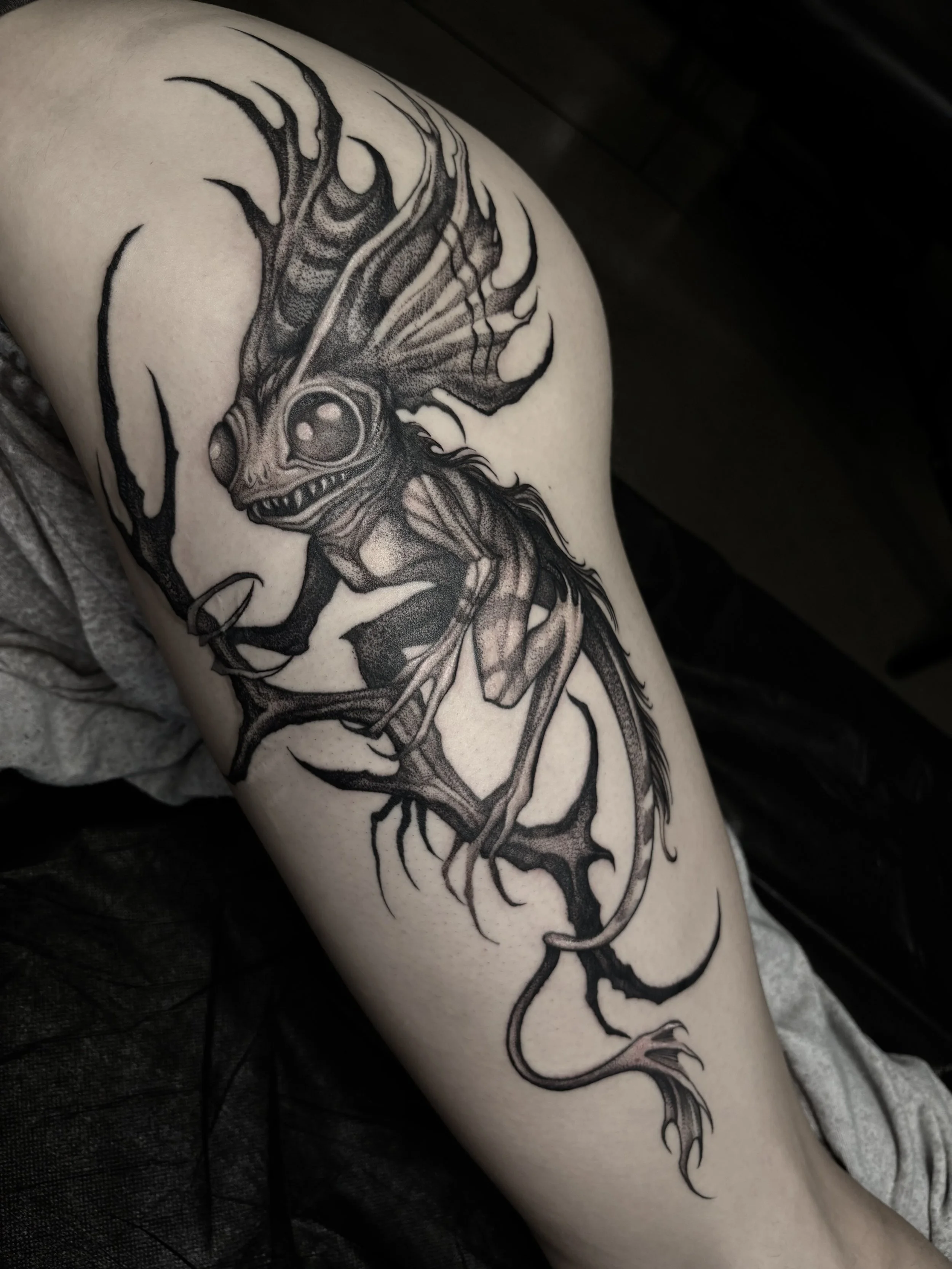 Black and gray tattoo of a mythical creature with a dragon-like head, large eyes, and a long, winding body with sharp, jagged features on the arm.