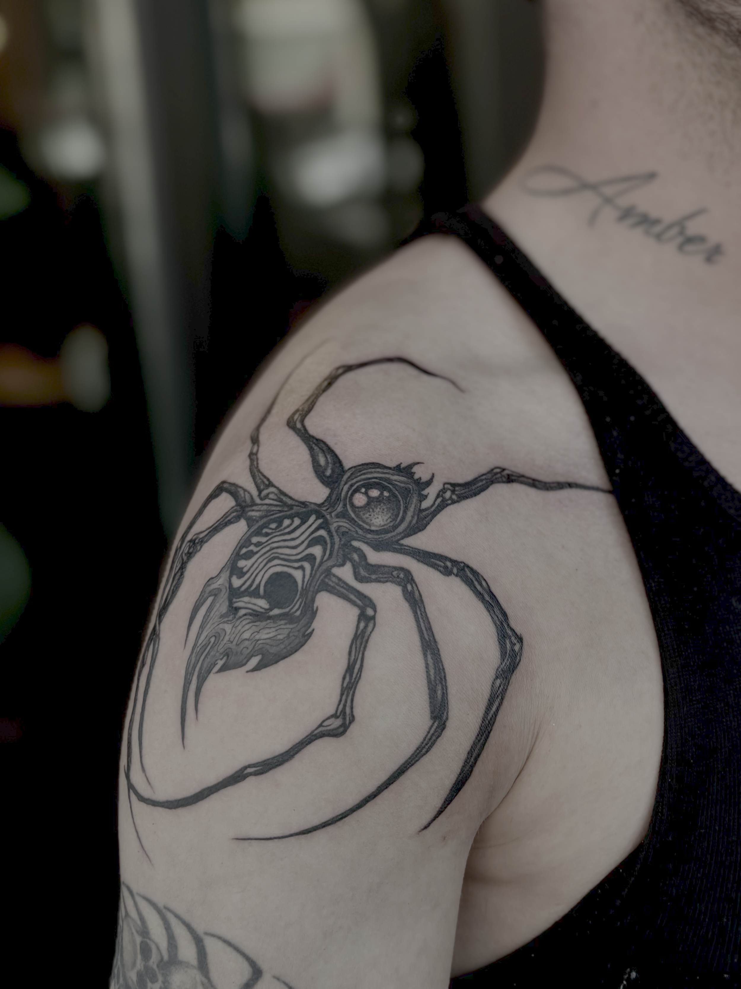 Close-up of a person's arm with a black tattoo of a spider and the word 'Ambe' written in cursive on the shoulder, wearing a black strap.