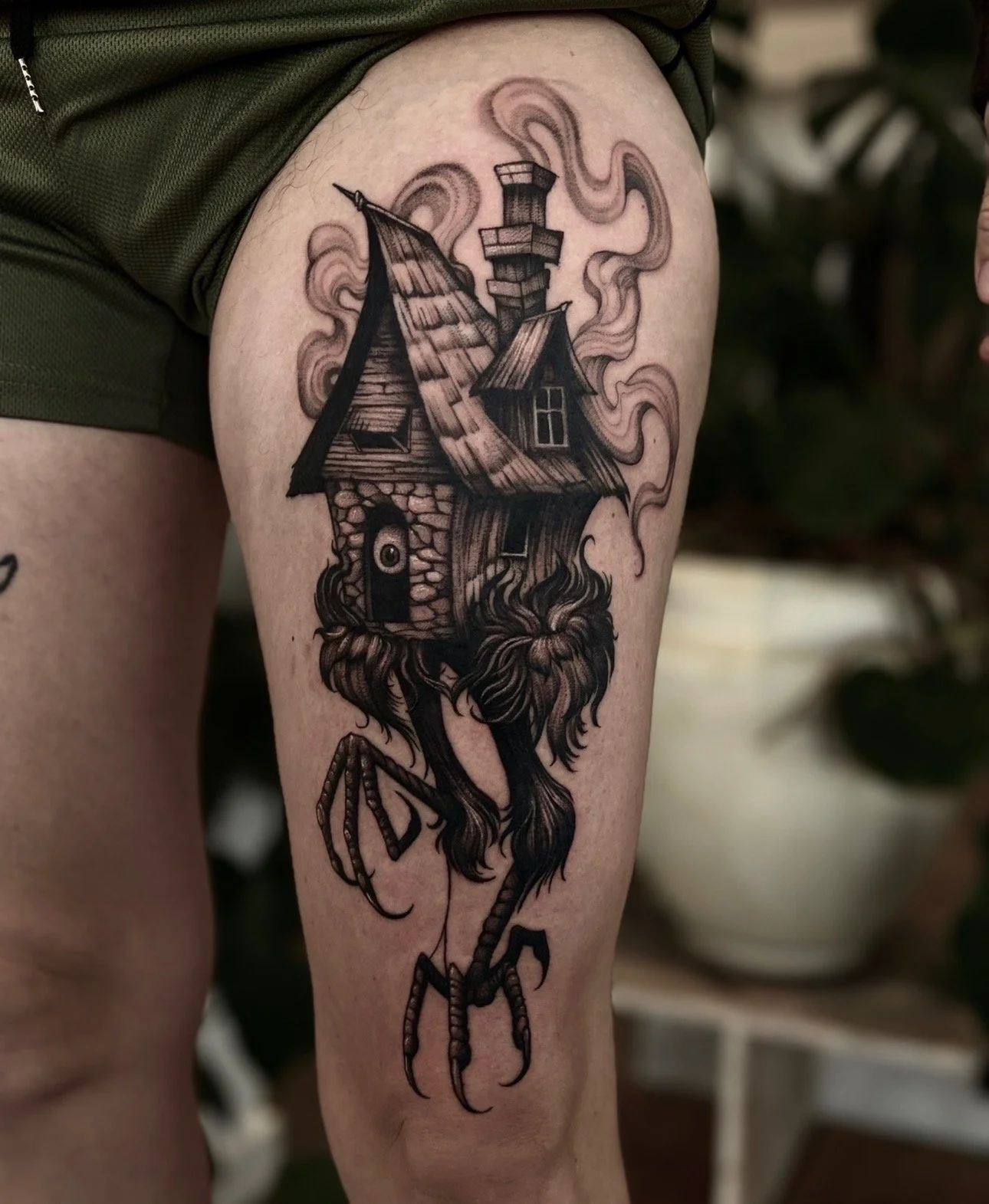 Tattoo of a house with a chimney, smoking, on a crab's back, with a claw and legs, on a person's thigh.