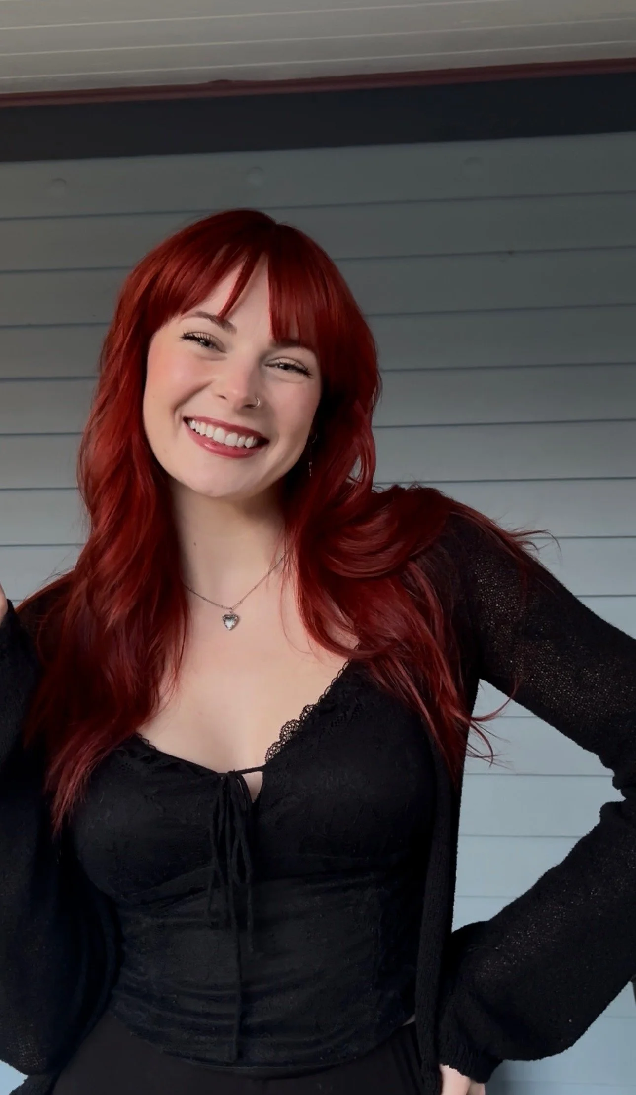 A young woman with red hair, smiling, wearing a black lace top, standing in front of a gray and black wall.
