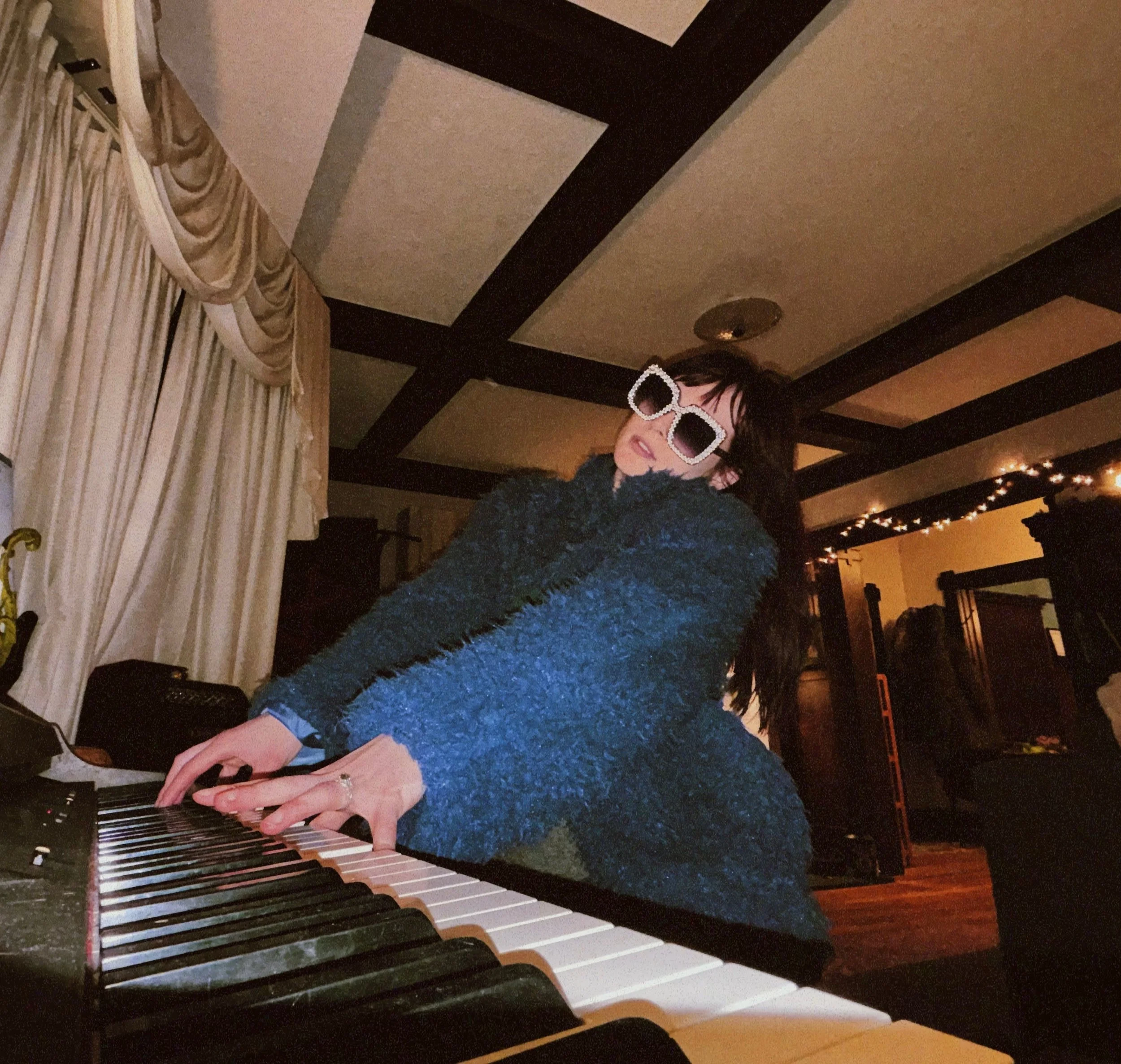 A woman wearing large white framed sunglasses playing a keyboard in a room with a dark wood and beige ceiling and beige curtains on the left.